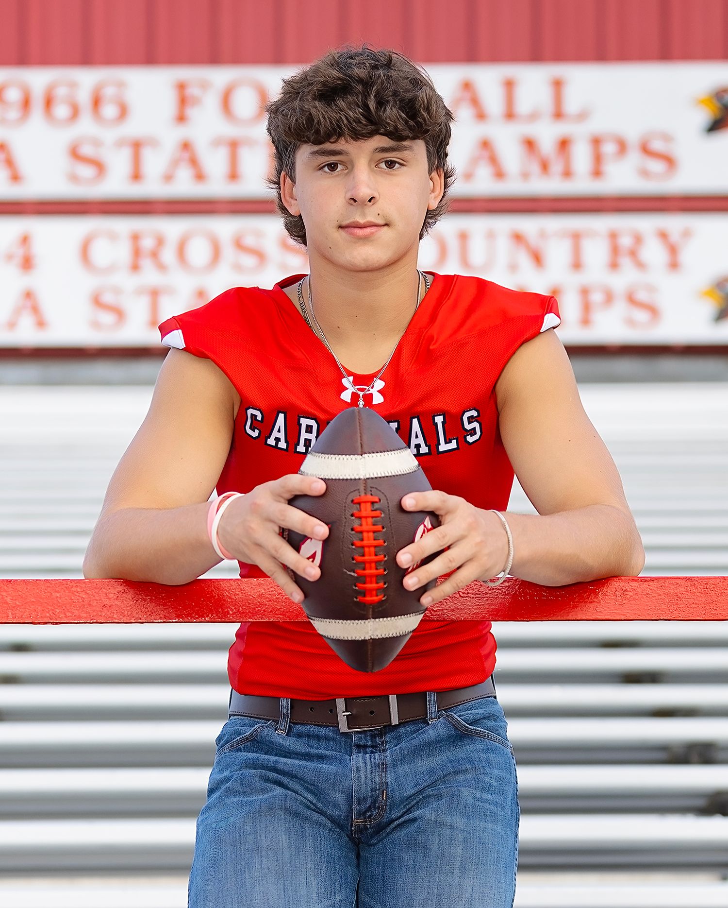 High school athlete in red Cardinals football jersey sits on bleachers holding football for sports portrait.