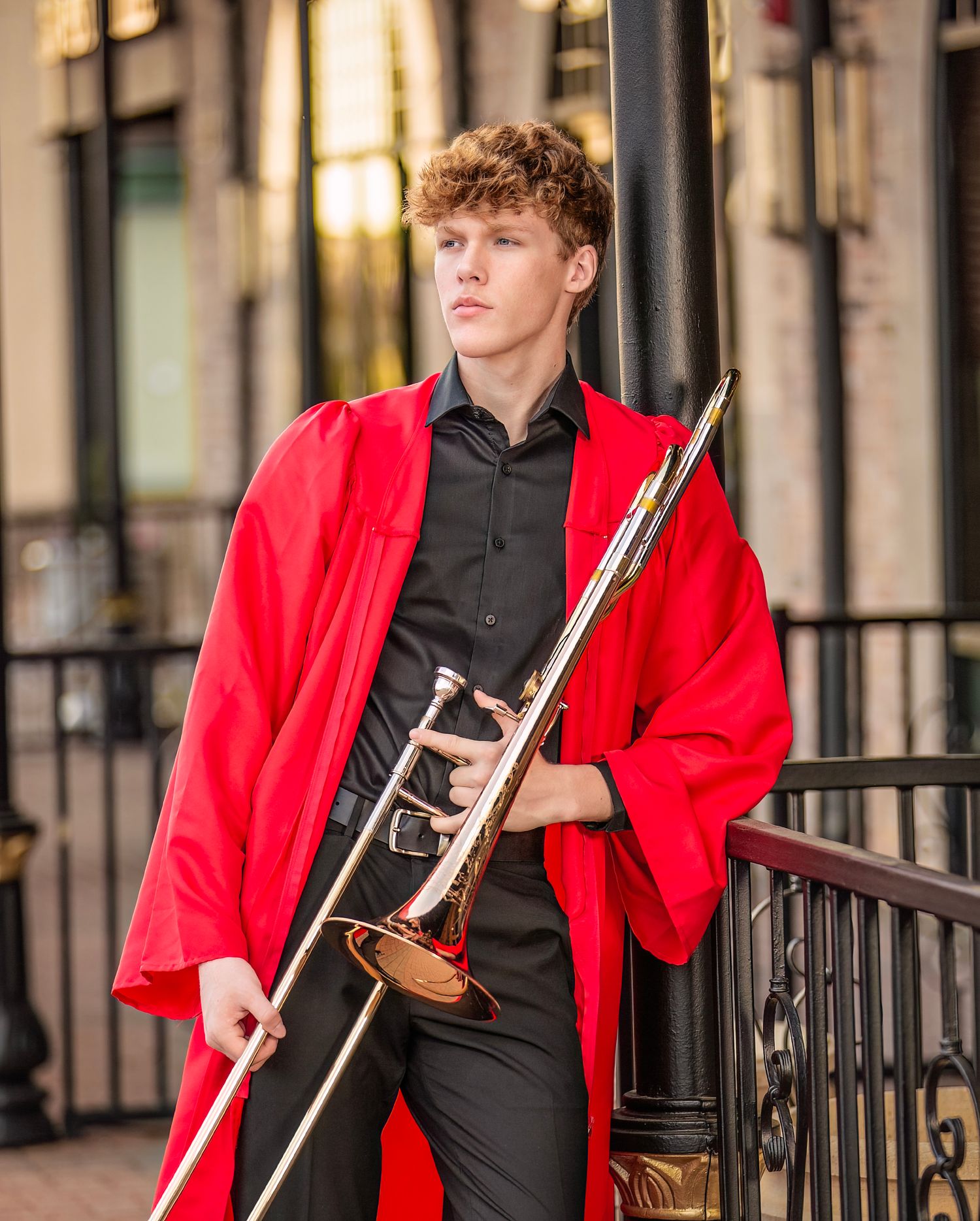Young male musician in red graduation gown holds a trombone while posing outdoors near iron railings.