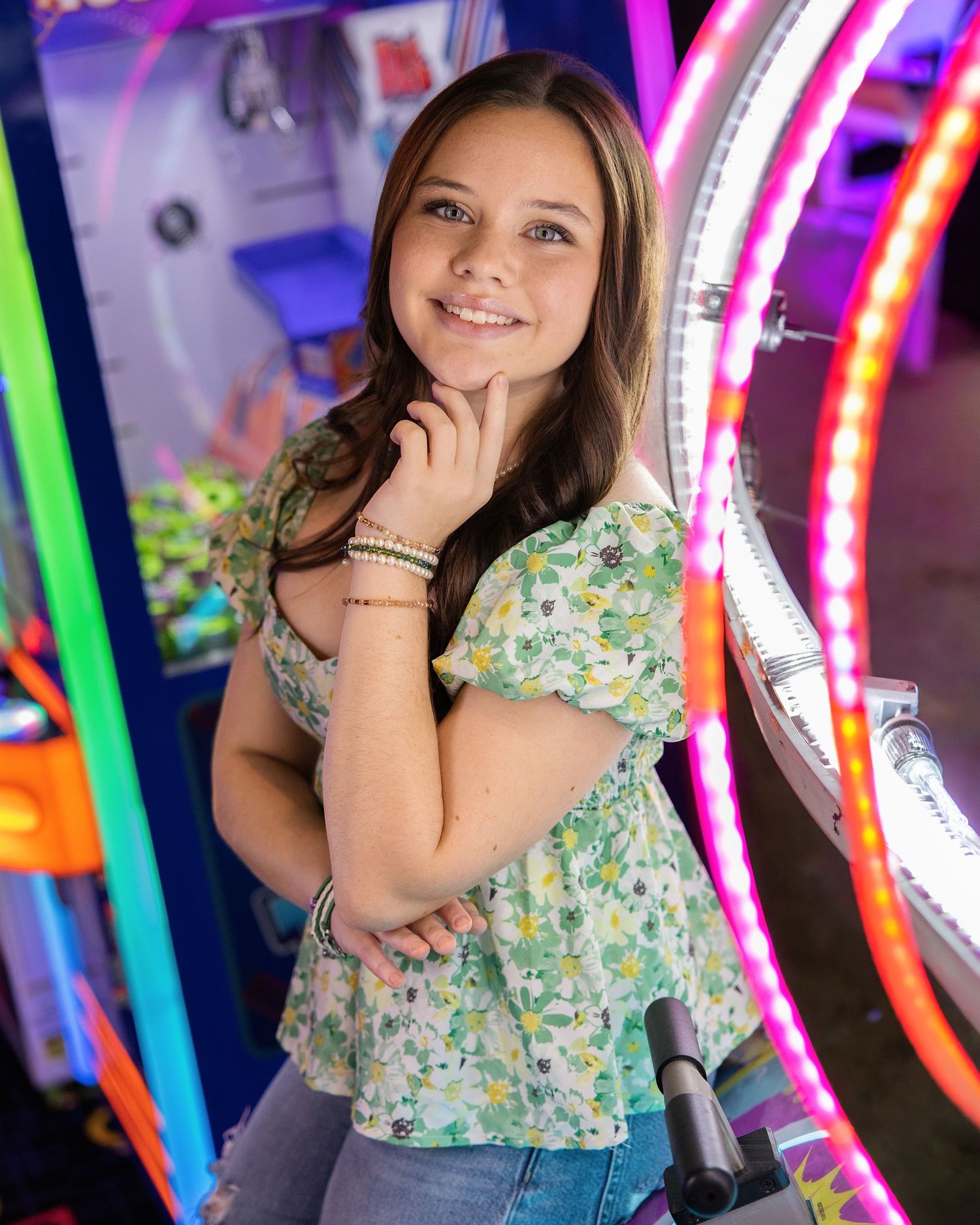 Young girl smiling in floral top at arcade with colorful neon lights in background.