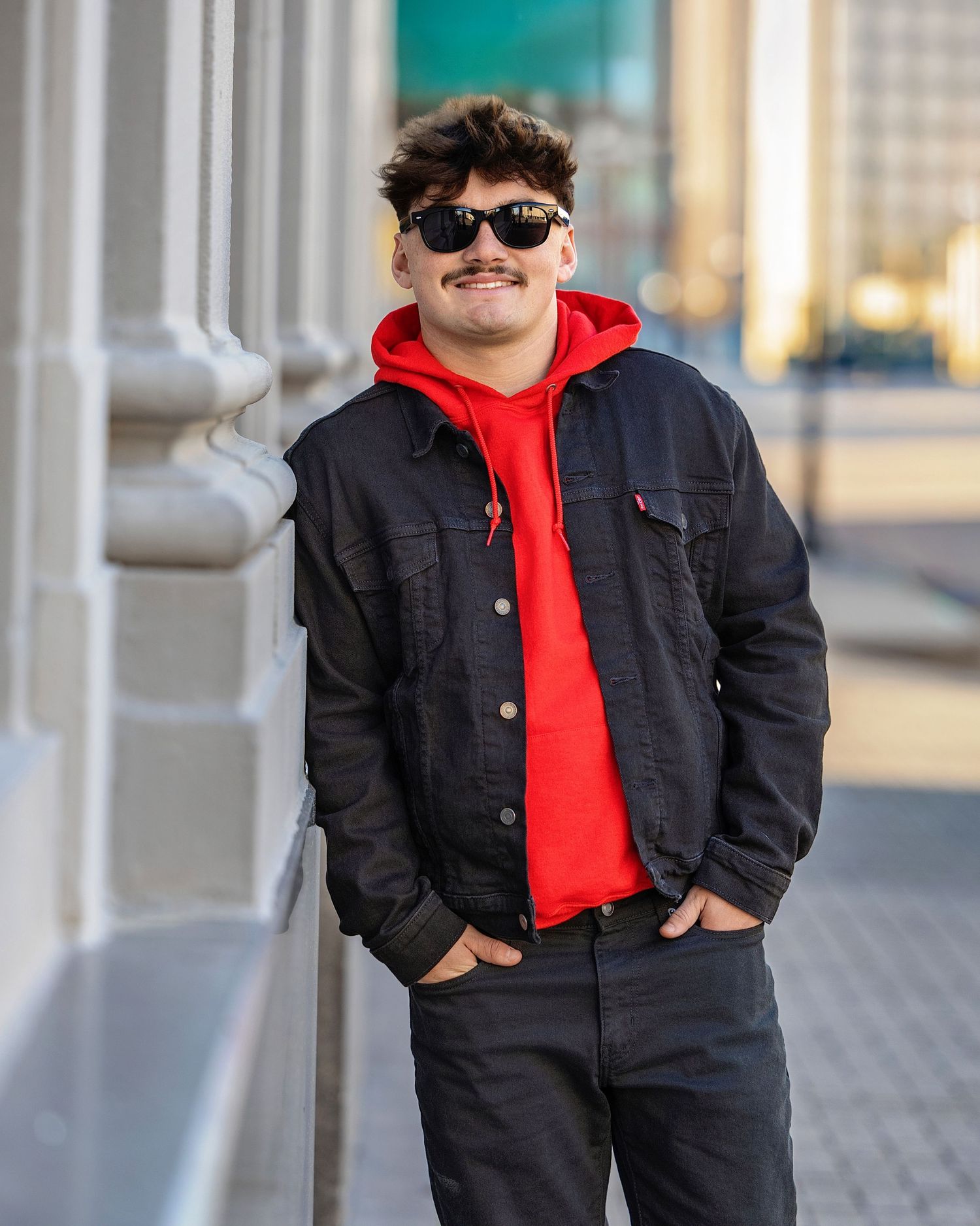 Young man with mustache wearing red hoodie, black denim jacket, and sunglasses leaning against urban building column.