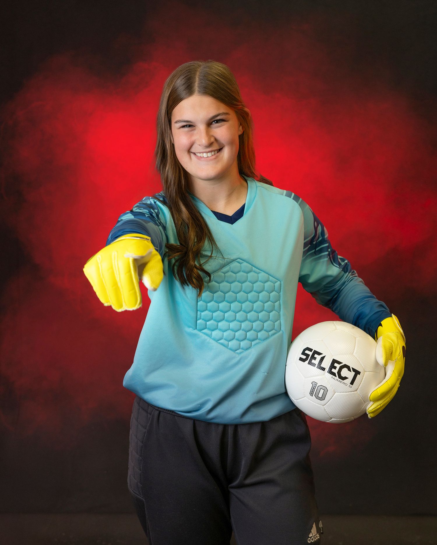Soccer goalkeeper in light blue jersey and yellow gloves holds a Select brand volleyball against dramatic red background.