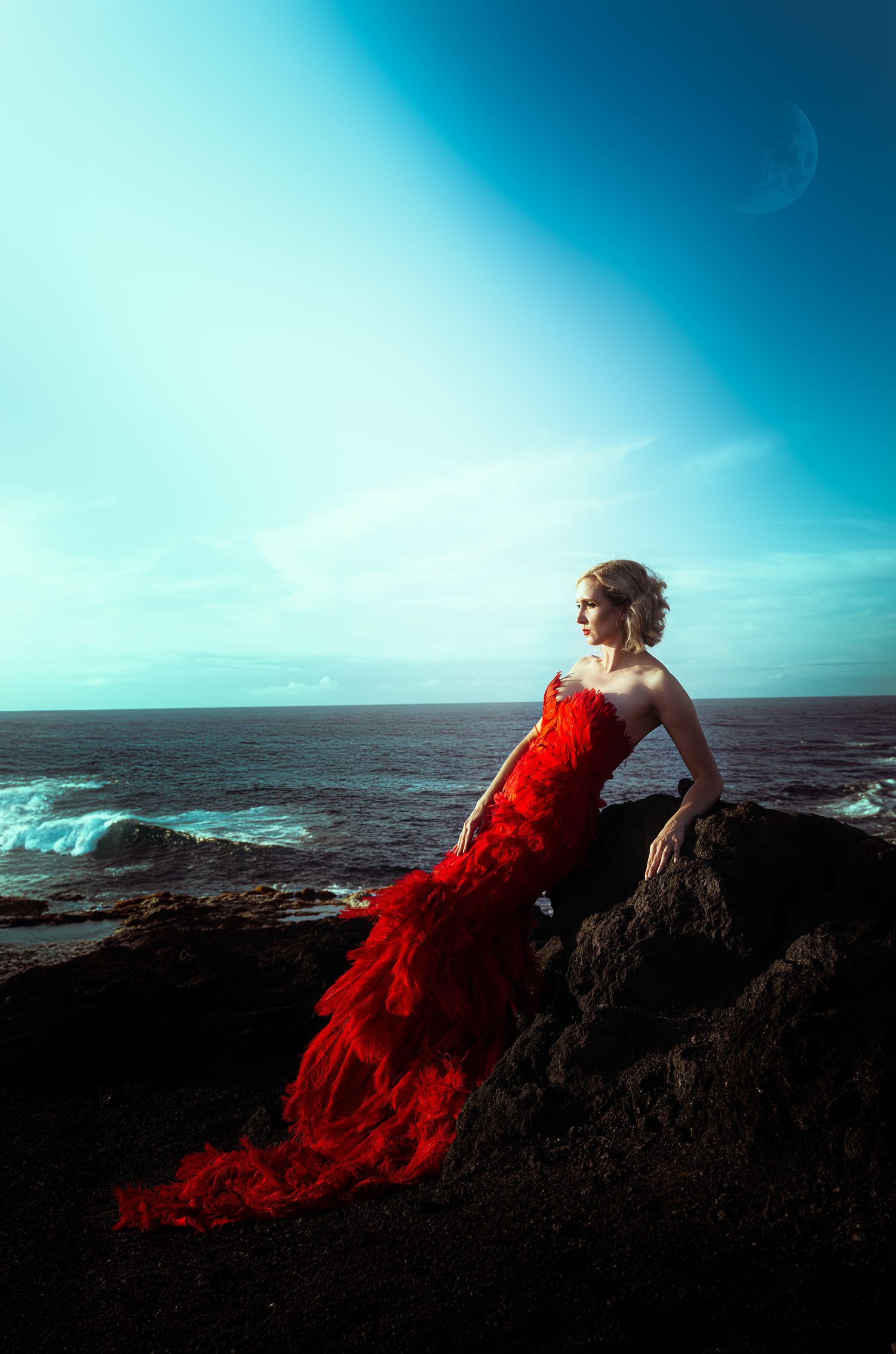 A woman in a stunning red mermaid gown sits on coastal rocks overlooking a vast turquoise ocean.
