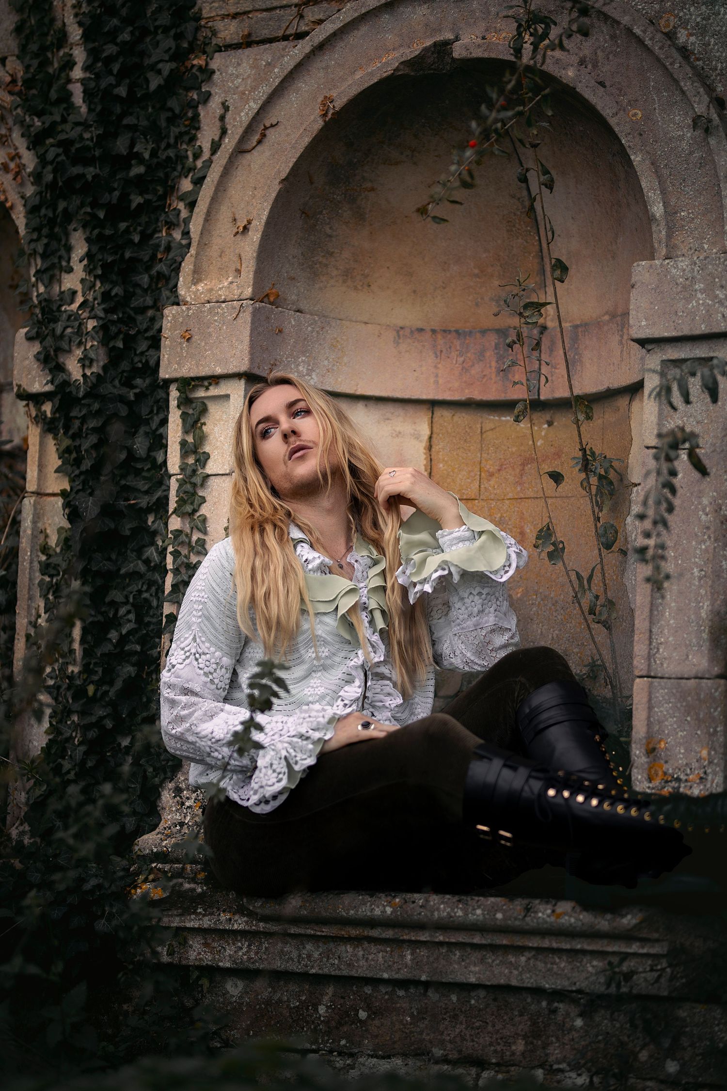 Blonde woman in vintage lace blouse sitting in ivy-covered stone niche of old ruins.