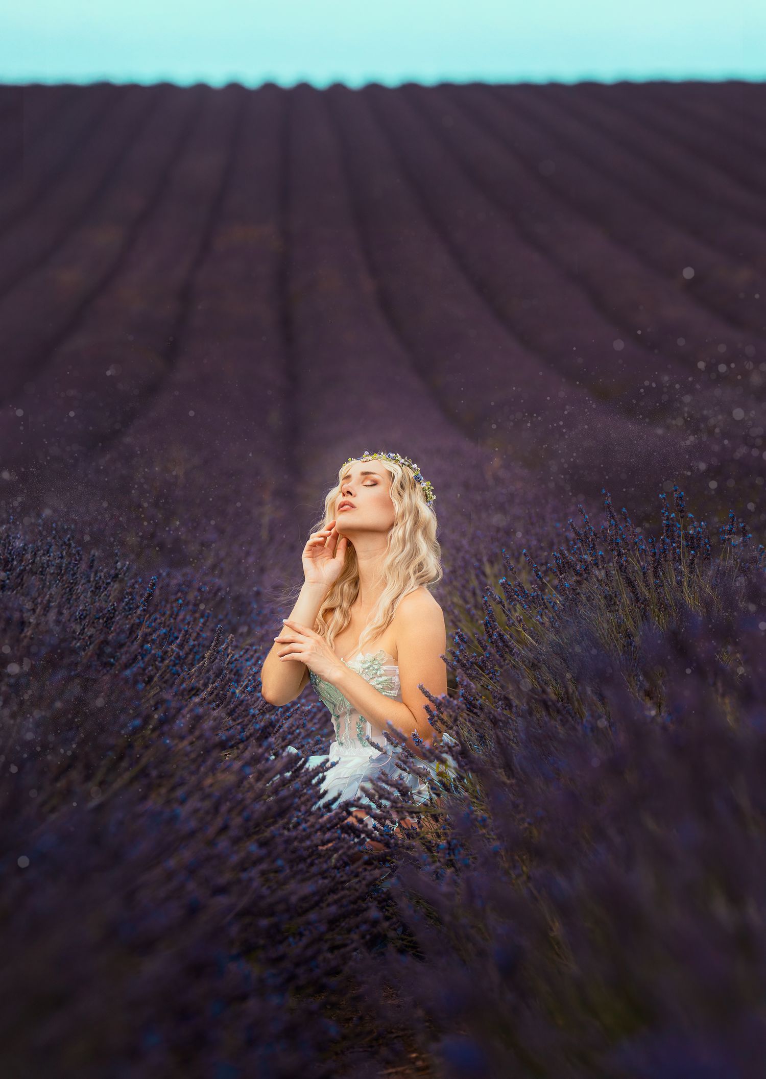 Blonde woman in white fairy dress with floral crown kneeling in vast lavender field.