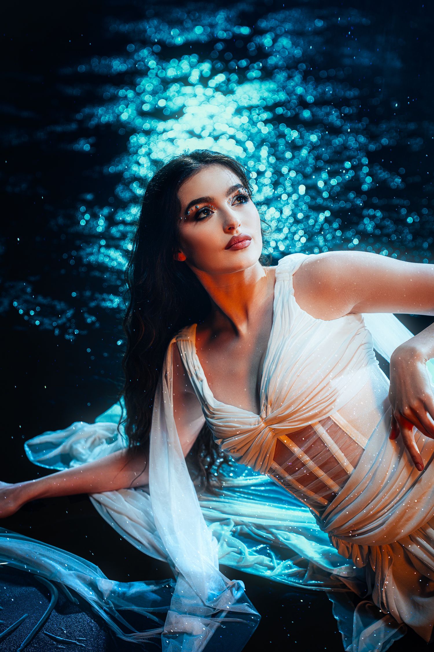 Young woman in white flowing dress poses gracefully against shimmering blue water backdrop at night.