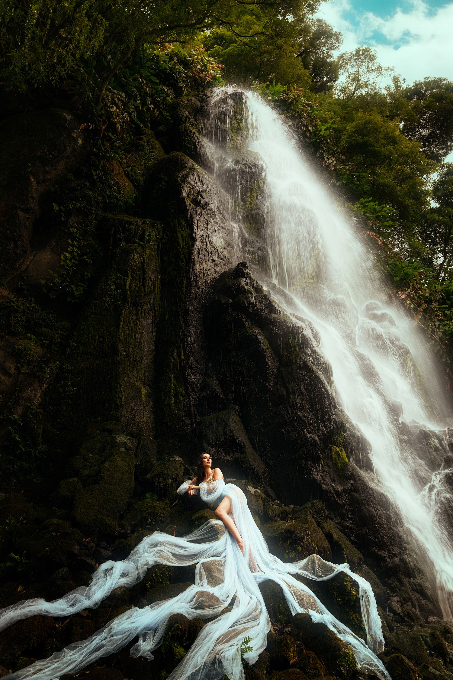 Woman in flowing white dress poses before a majestic waterfall surrounded by lush green forest.