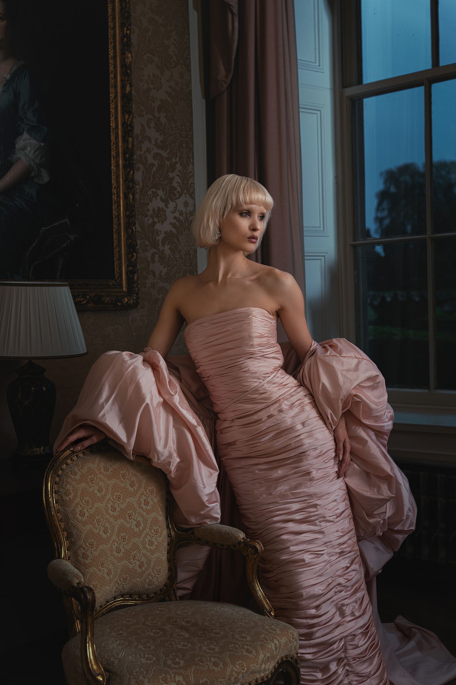 A blonde woman in a strapless blush pink ruched gown sits elegantly in a vintage armchair in a classic interior.