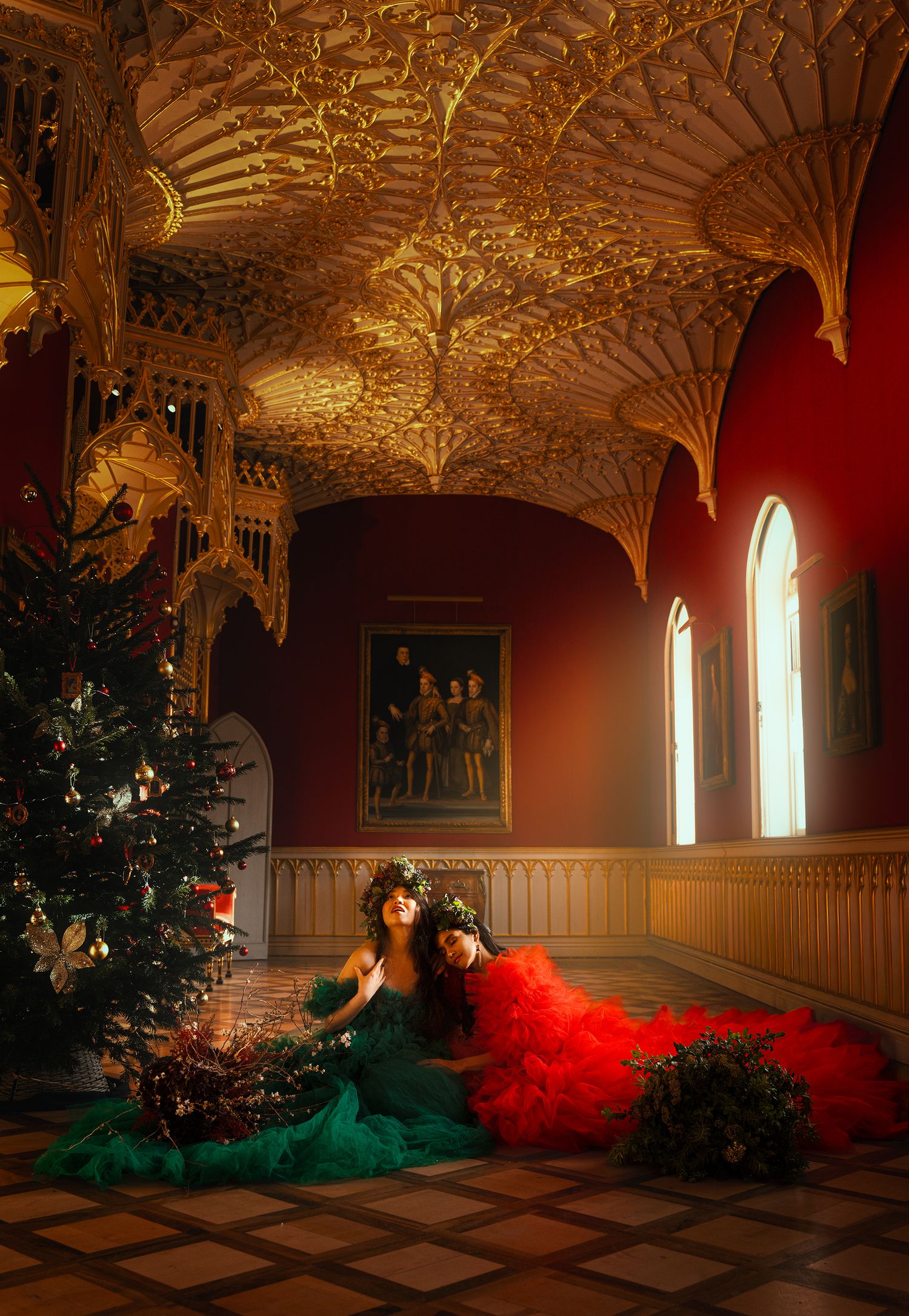 Two women in red and green ball gowns resting beside Christmas tree in grand Gothic hall.