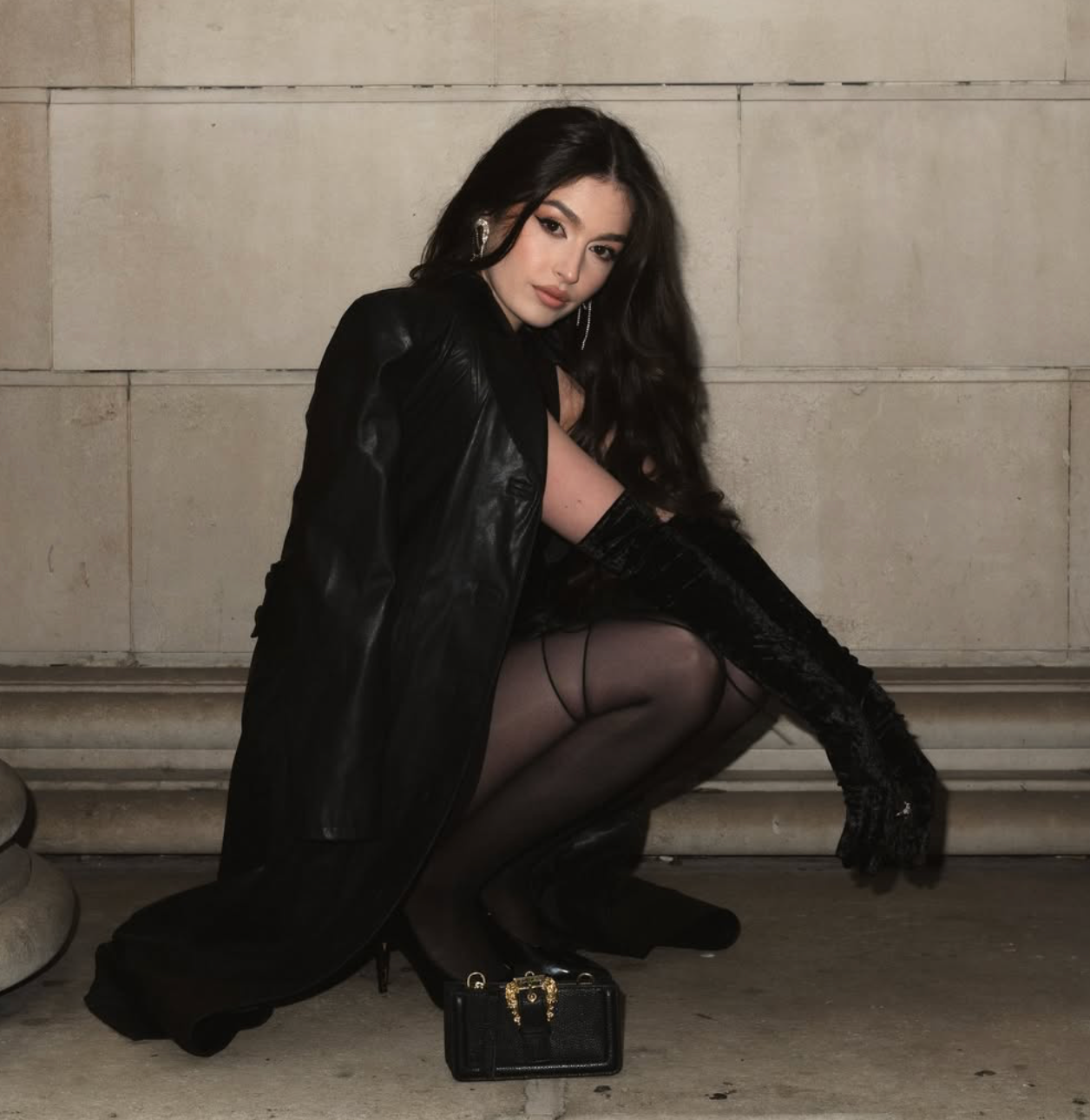 Stylish woman in black fur coat and sheer tights crouching with luxury handbag in urban setting.