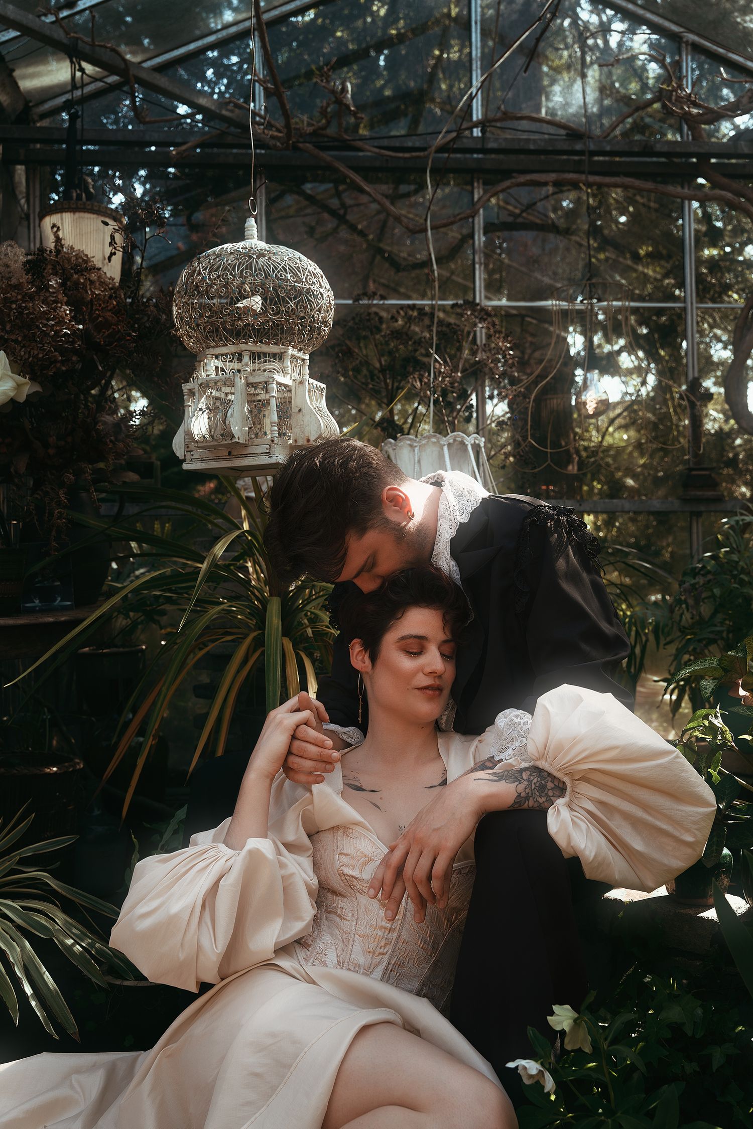A couple in Victorian attire share a romantic moment in a lush greenhouse setting.