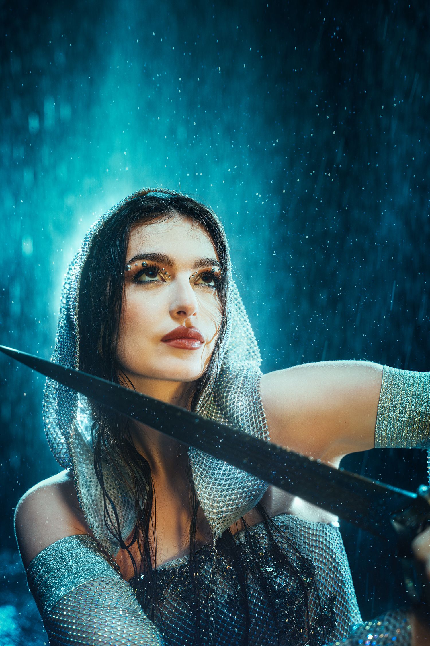 Warrior woman in silver mesh hood holding crossed swords in blue rain.