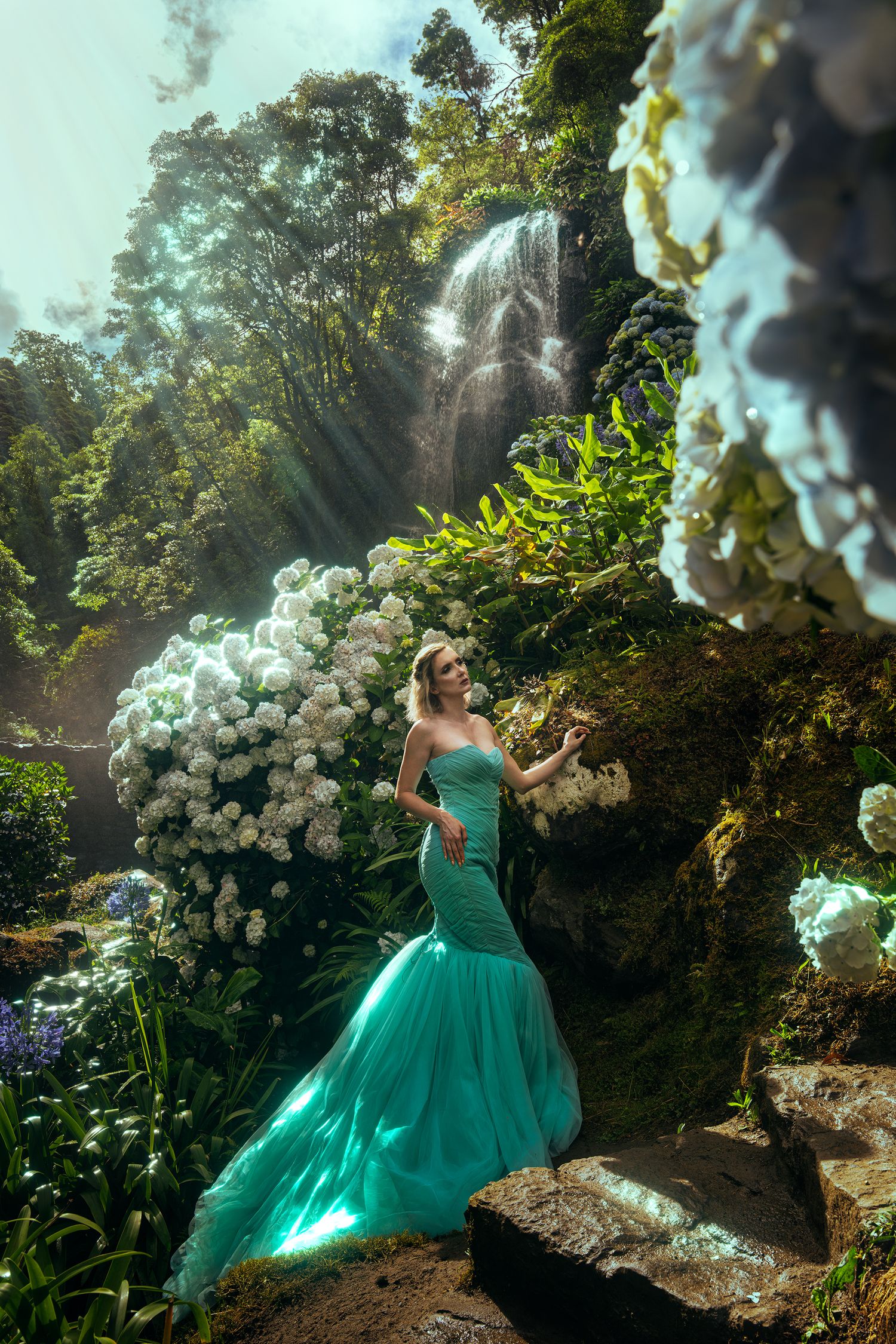 Blonde woman in teal mermaid gown stands among white flowers near a lush forest waterfall.