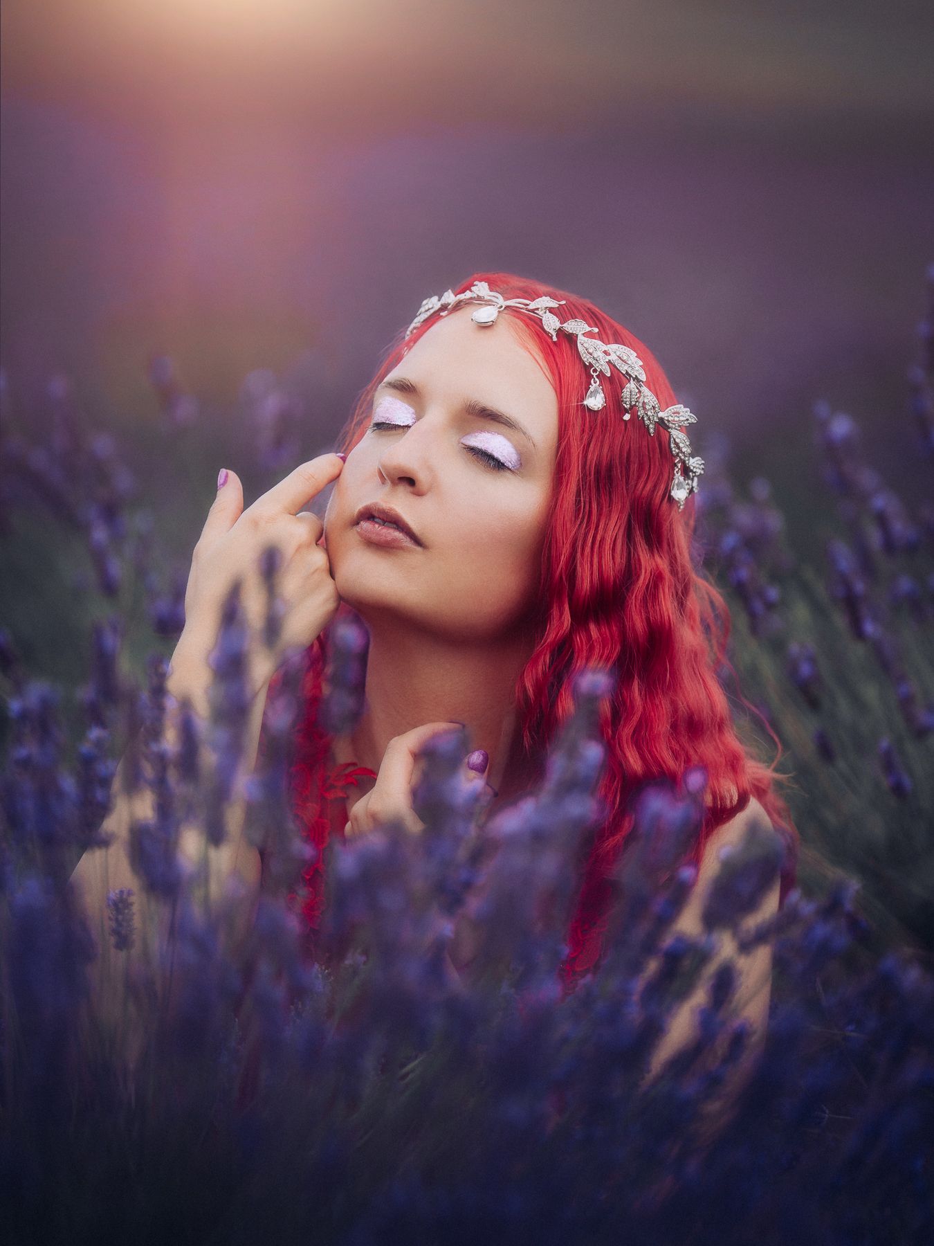Woman with red hair wearing silver floral crown poses dreamily in a purple lavender field at sunset.