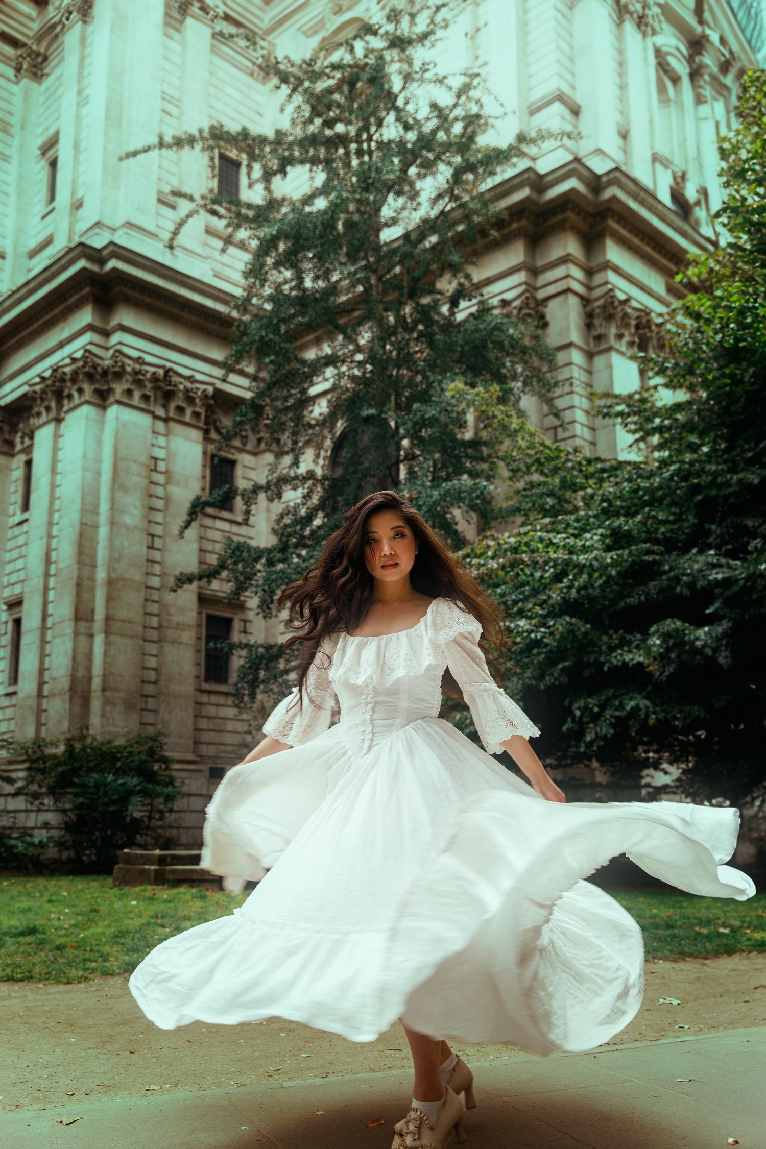 A woman in a flowing white dress twirls outdoors in front of a grand historic building surrounded by lush green trees.