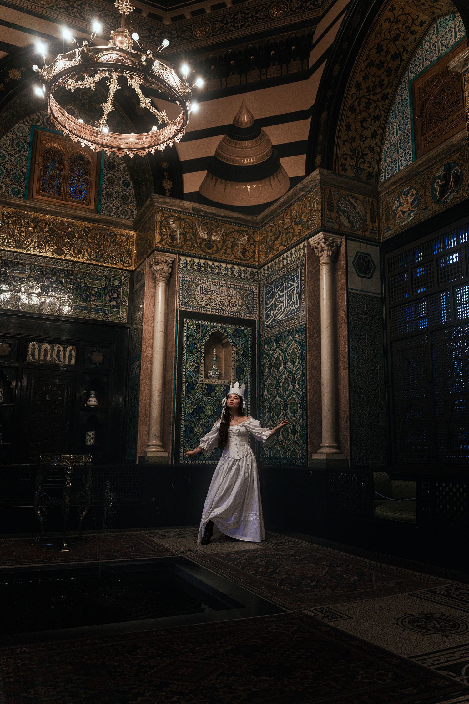 Woman in white gown and crown dancing in ornate Moorish-style tiled interior hall.