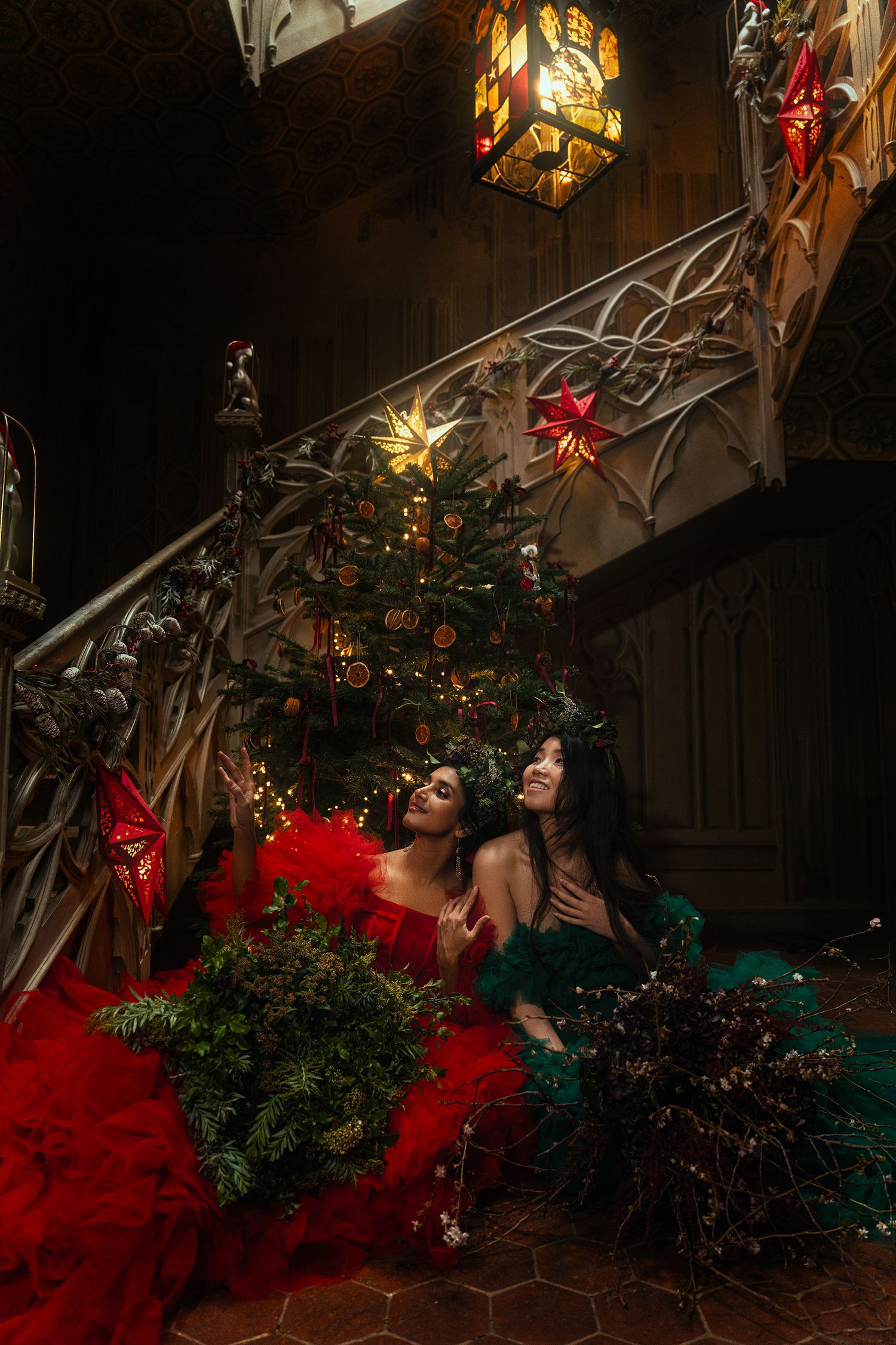 Two women in red and green ballgowns laugh together beside Christmas tree on decorated Gothic staircase.