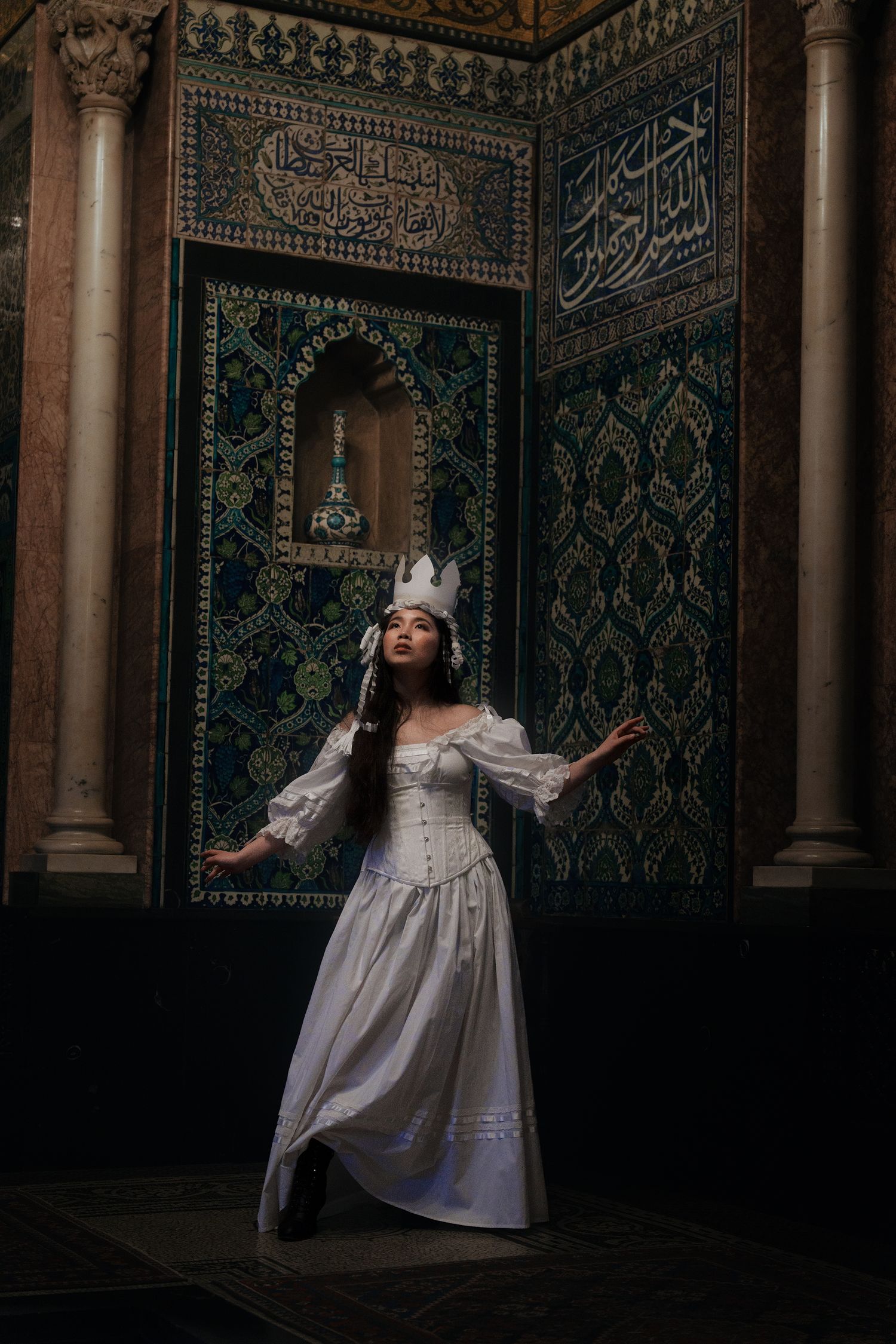Woman in white Victorian dress and paper crown poses against intricate Islamic tilework and Arabic calligraphy in historic interior.
