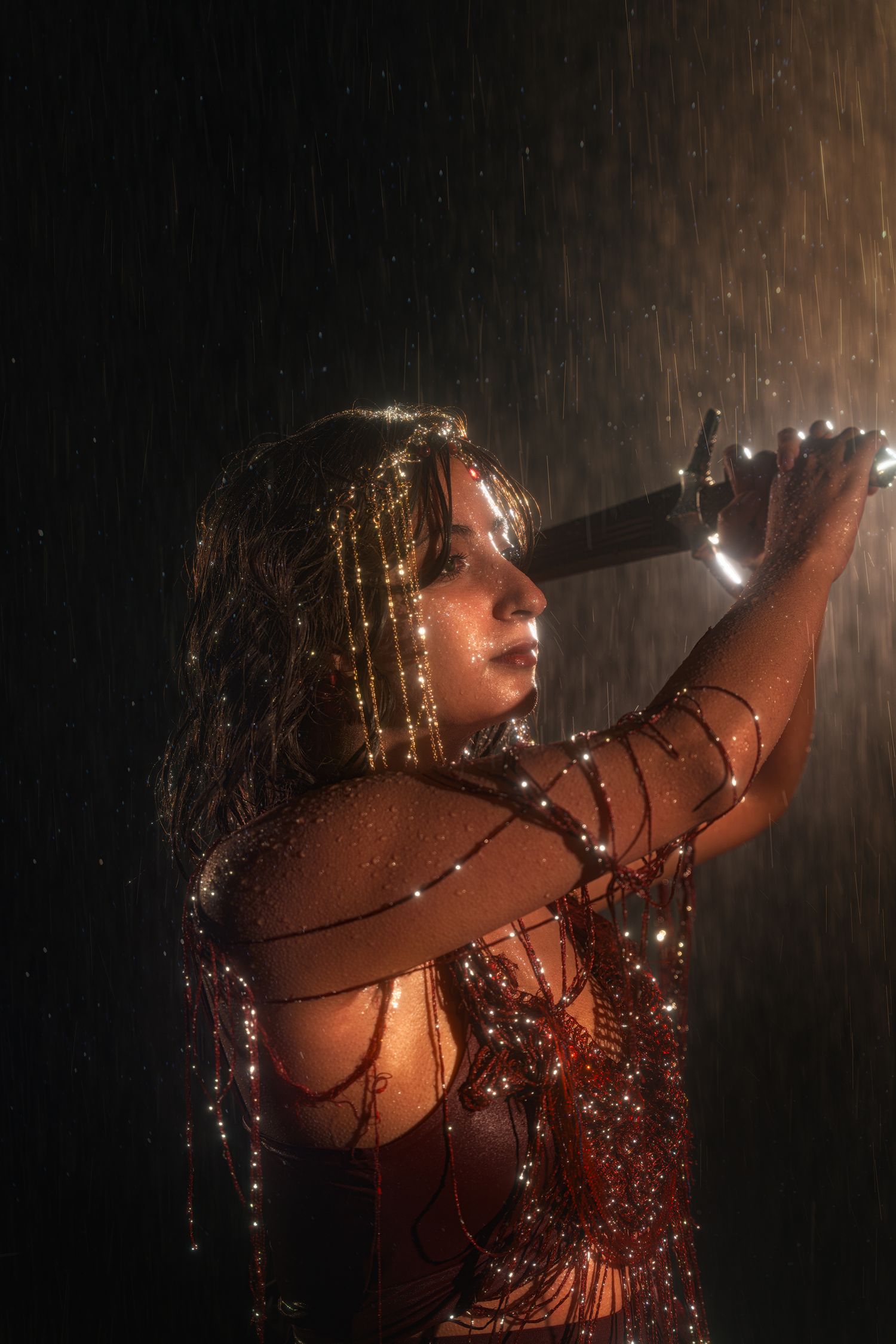 Woman in red outfit holds glowing sword aloft in dramatic rain shower with warm light.