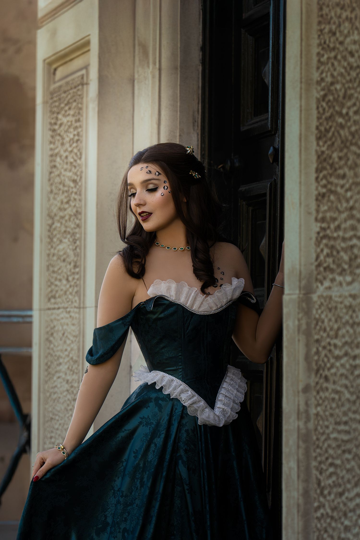Woman in dark teal Victorian corset gown with dramatic makeup leans against ornate building doorway.