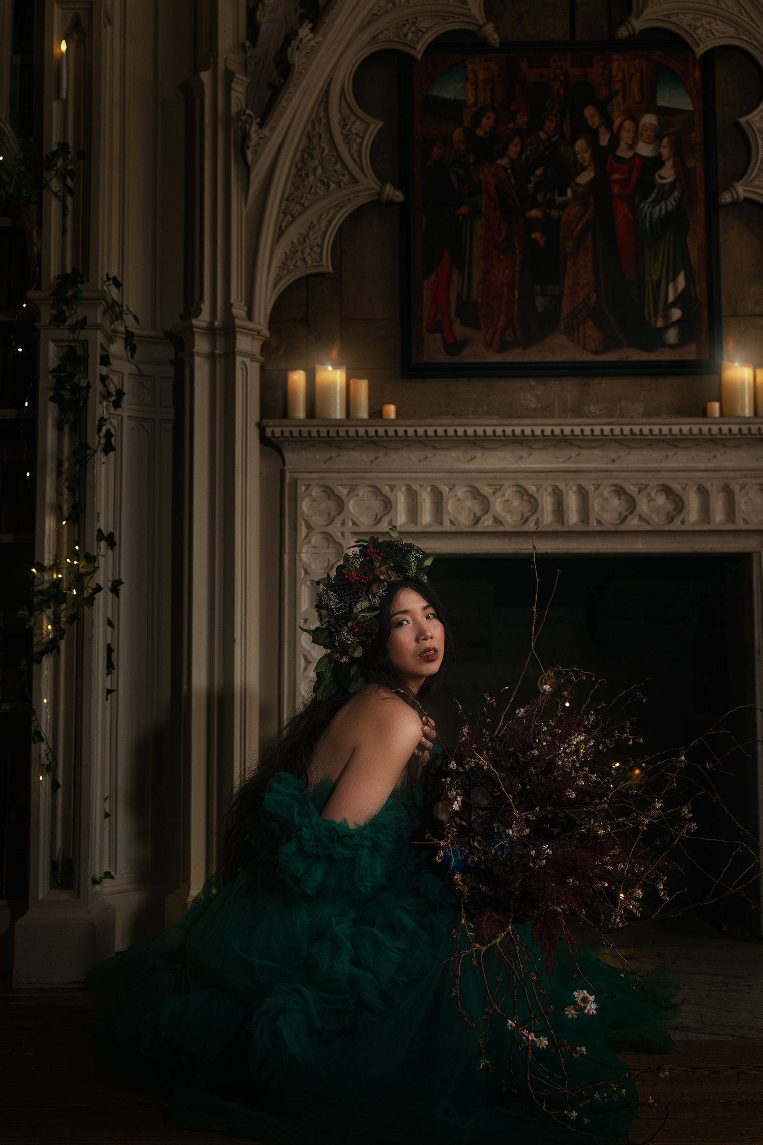 Woman in green tulle ballgown with floral crown sits pensively before Gothic candlelit fireplace.