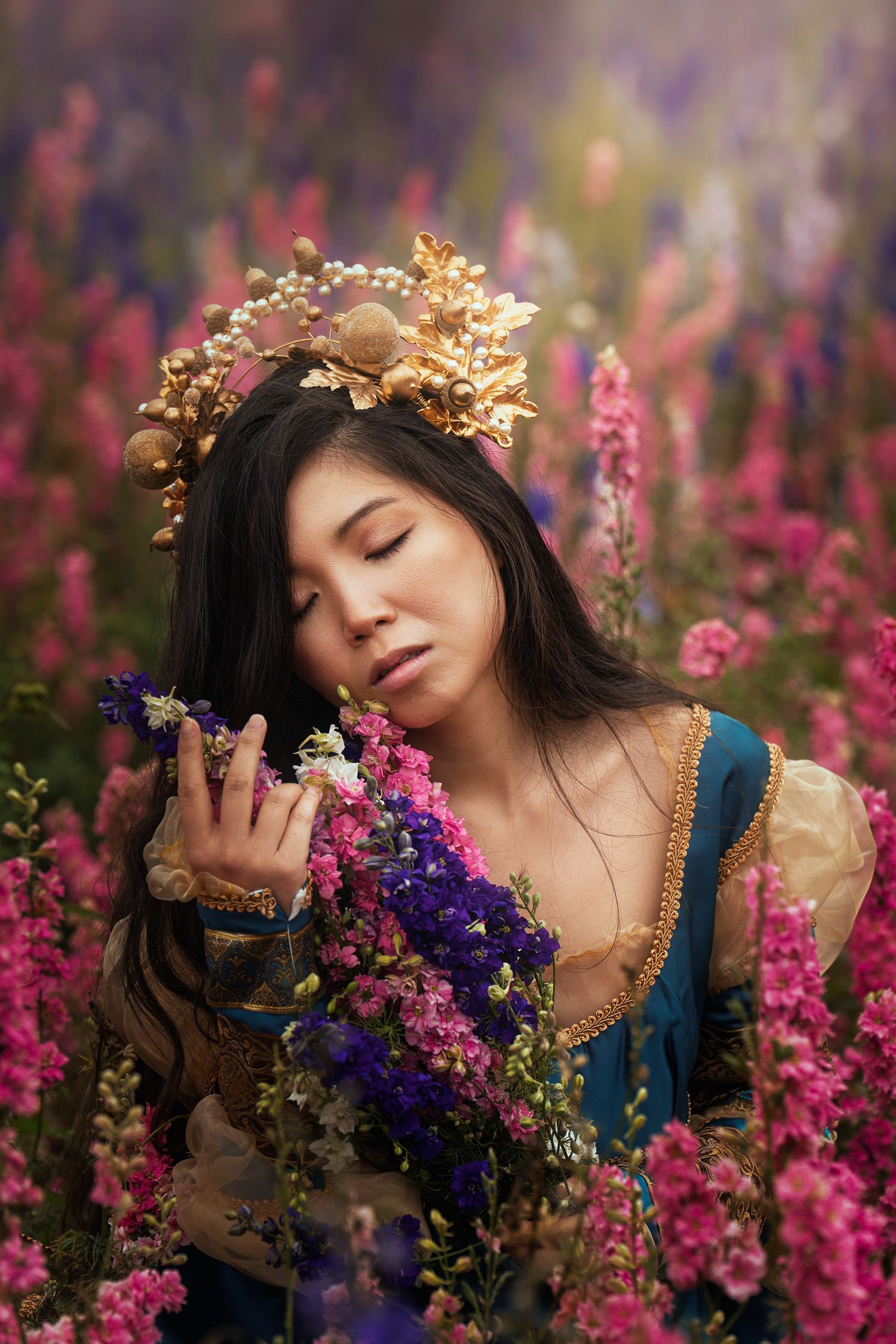 A woman wearing a gold crown and medieval blue dress holds flowers in a vibrant pink flower field.