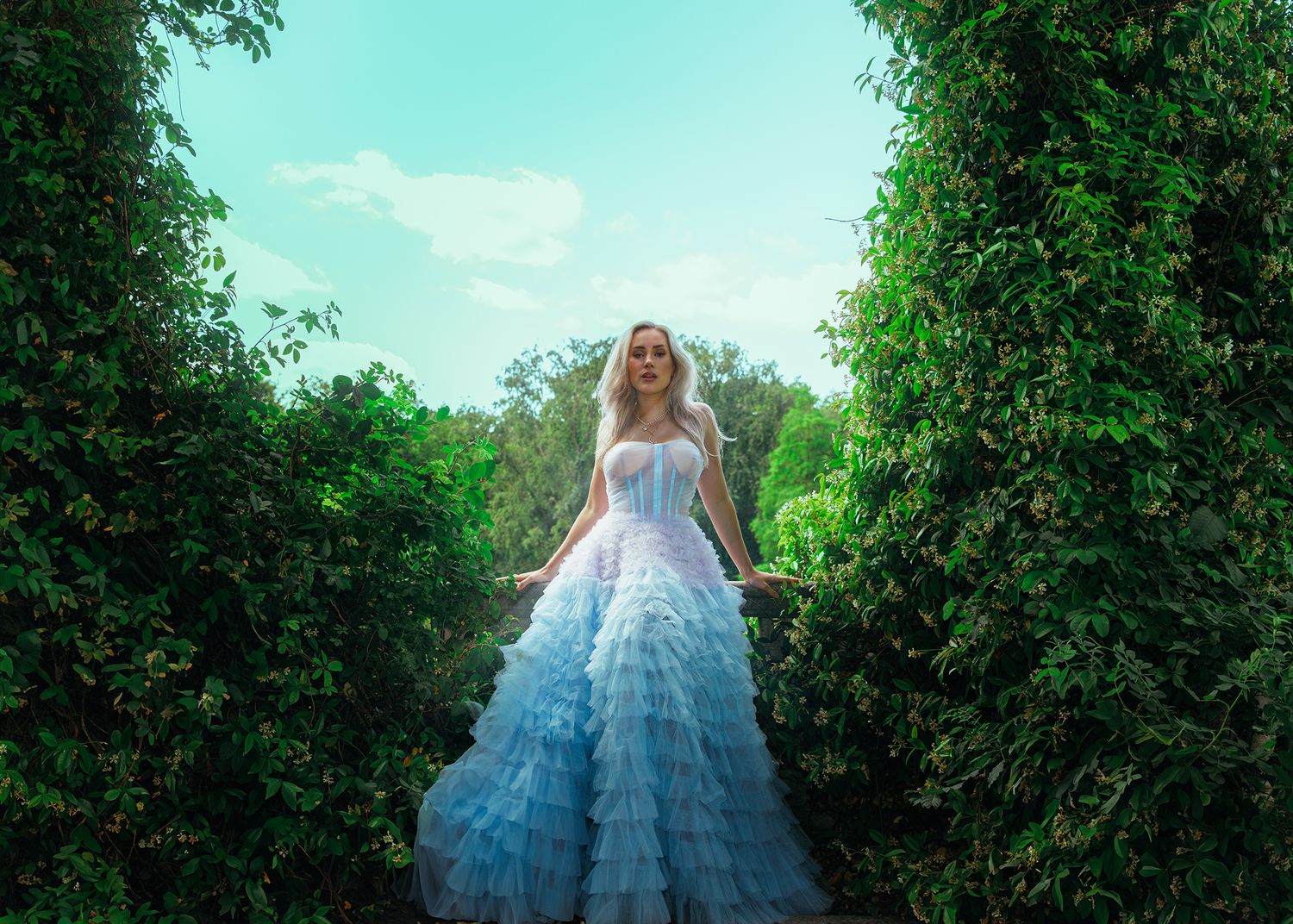 Blonde woman in layered light blue ball gown stands between tall green hedges under bright blue sky.