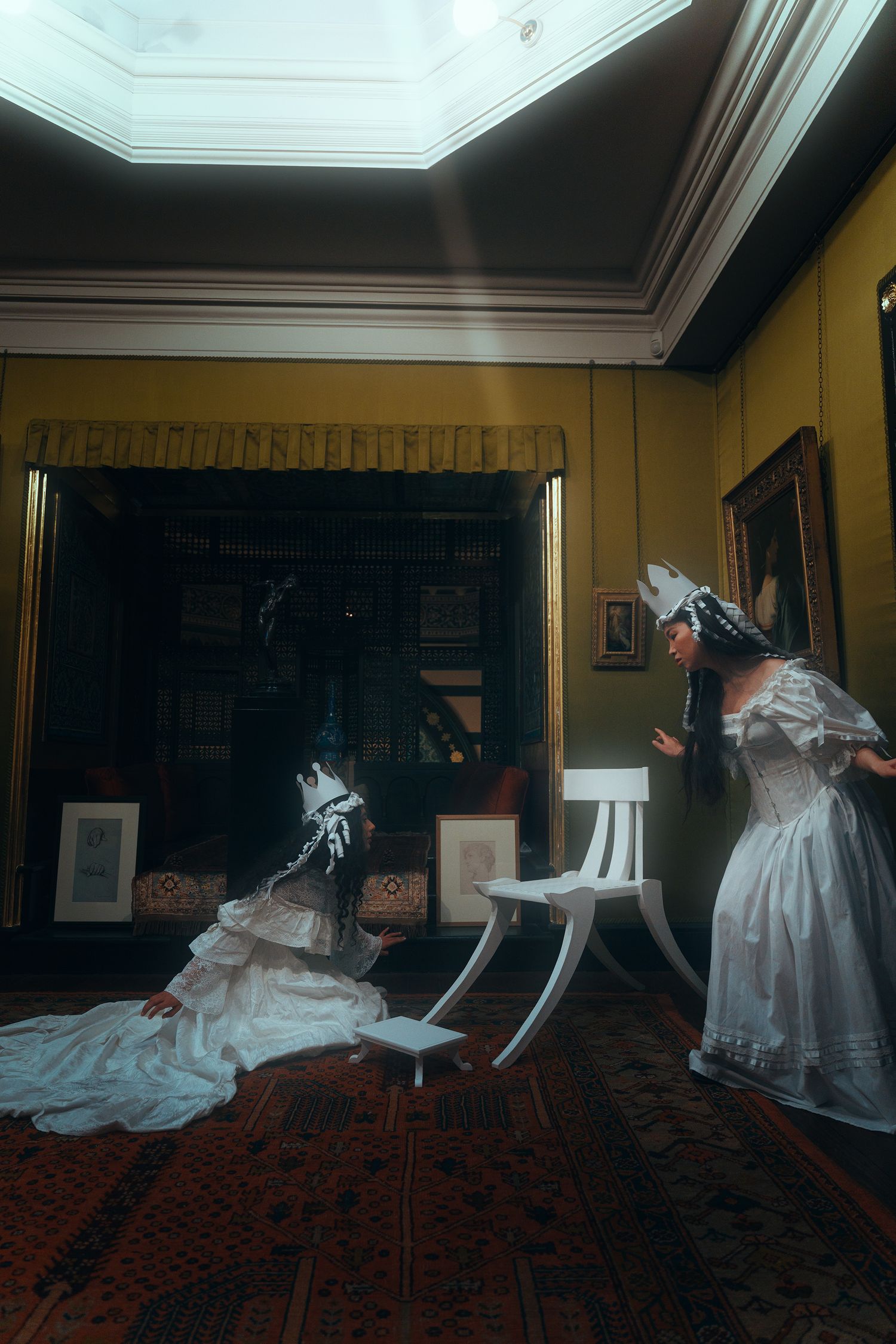 Two performers in white period dresses and decorative headpieces interact near a white chair in a dimly lit historic room.