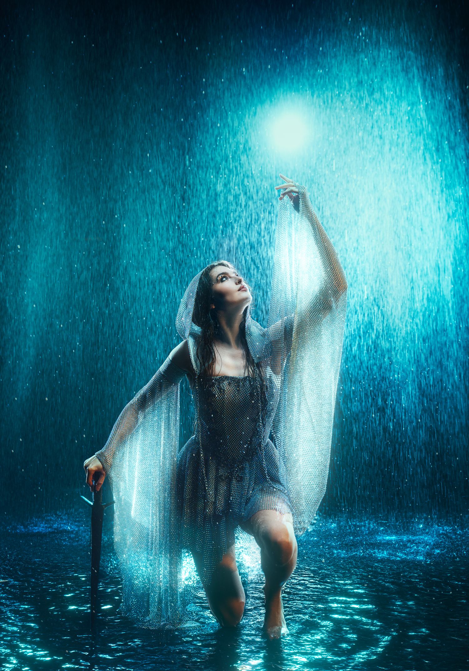 Fantasy woman in silver gown reaching for glowing orb while wading in rain.