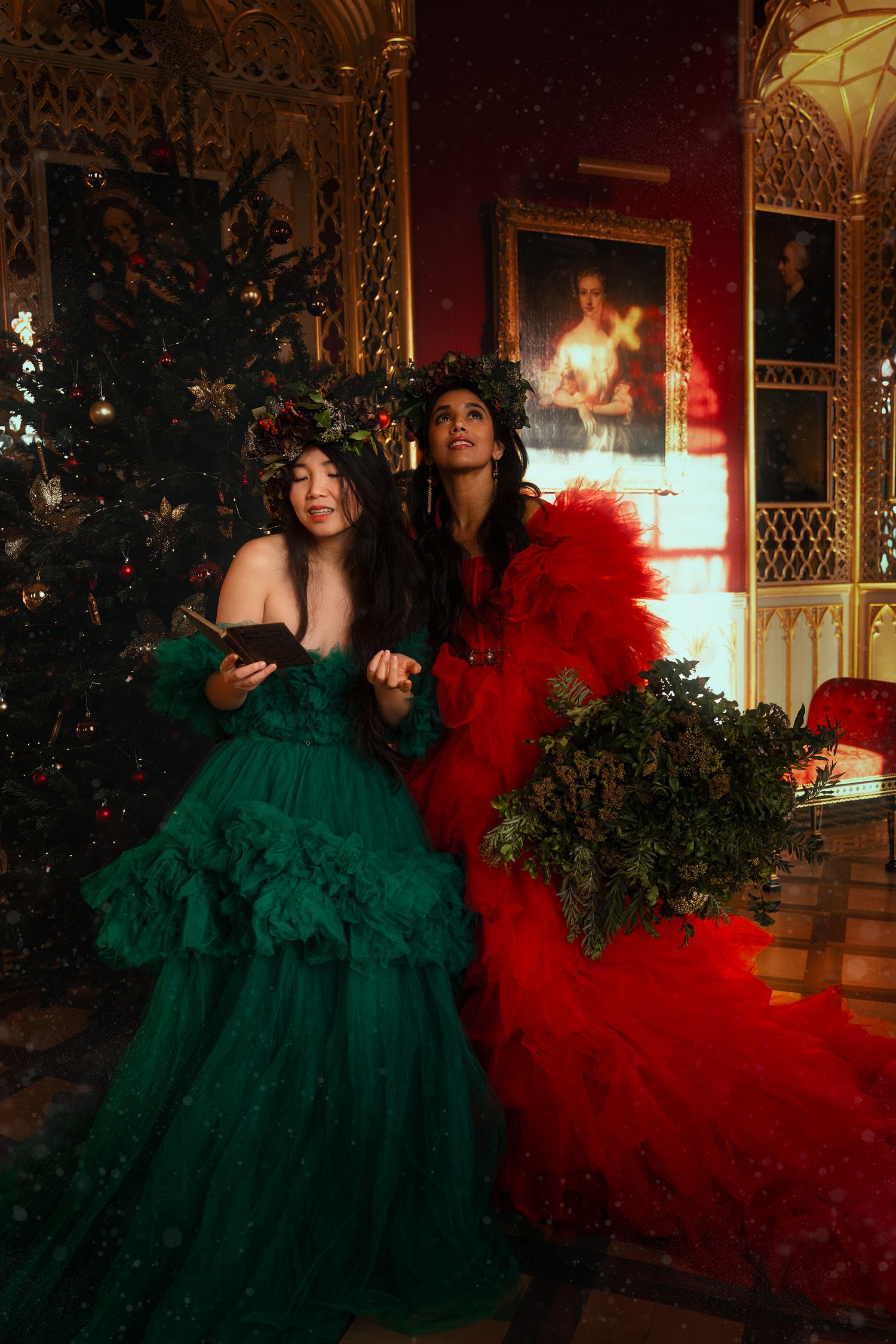 Two women in green and red ball gowns with floral crowns beside a Christmas tree in ornate hall.