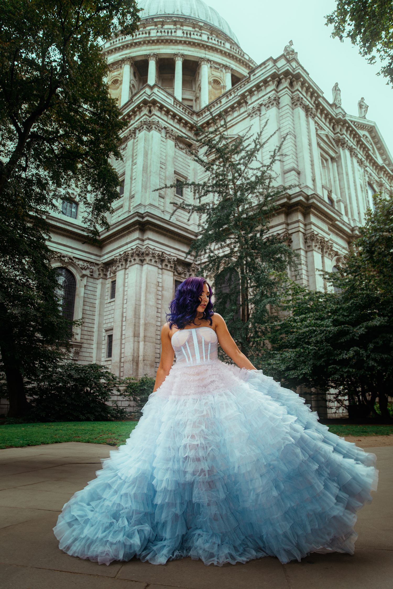 A woman with purple hair wears a stunning blue ombre ruffled ball gown in front of a grand domed cathedral.