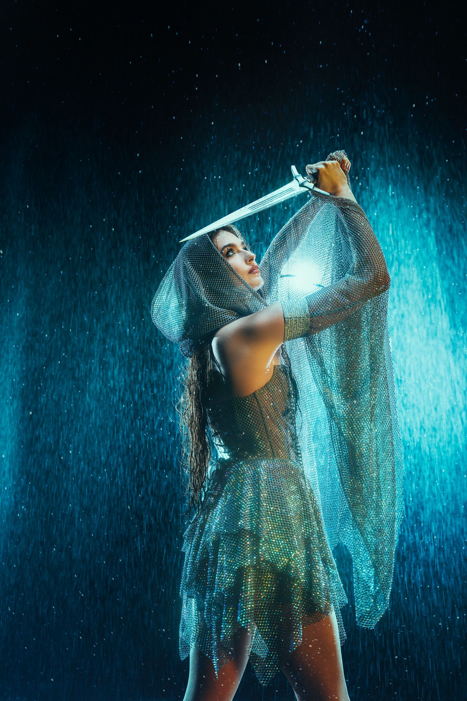 Fantasy warrior woman in sheer silver dress raising sword in dramatic rain storm.
