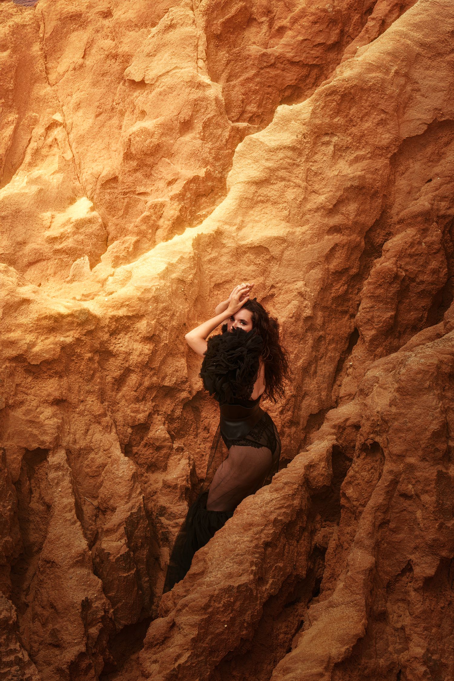 Woman in black outfit leans against golden sandstone rock formation in dramatic warm light.