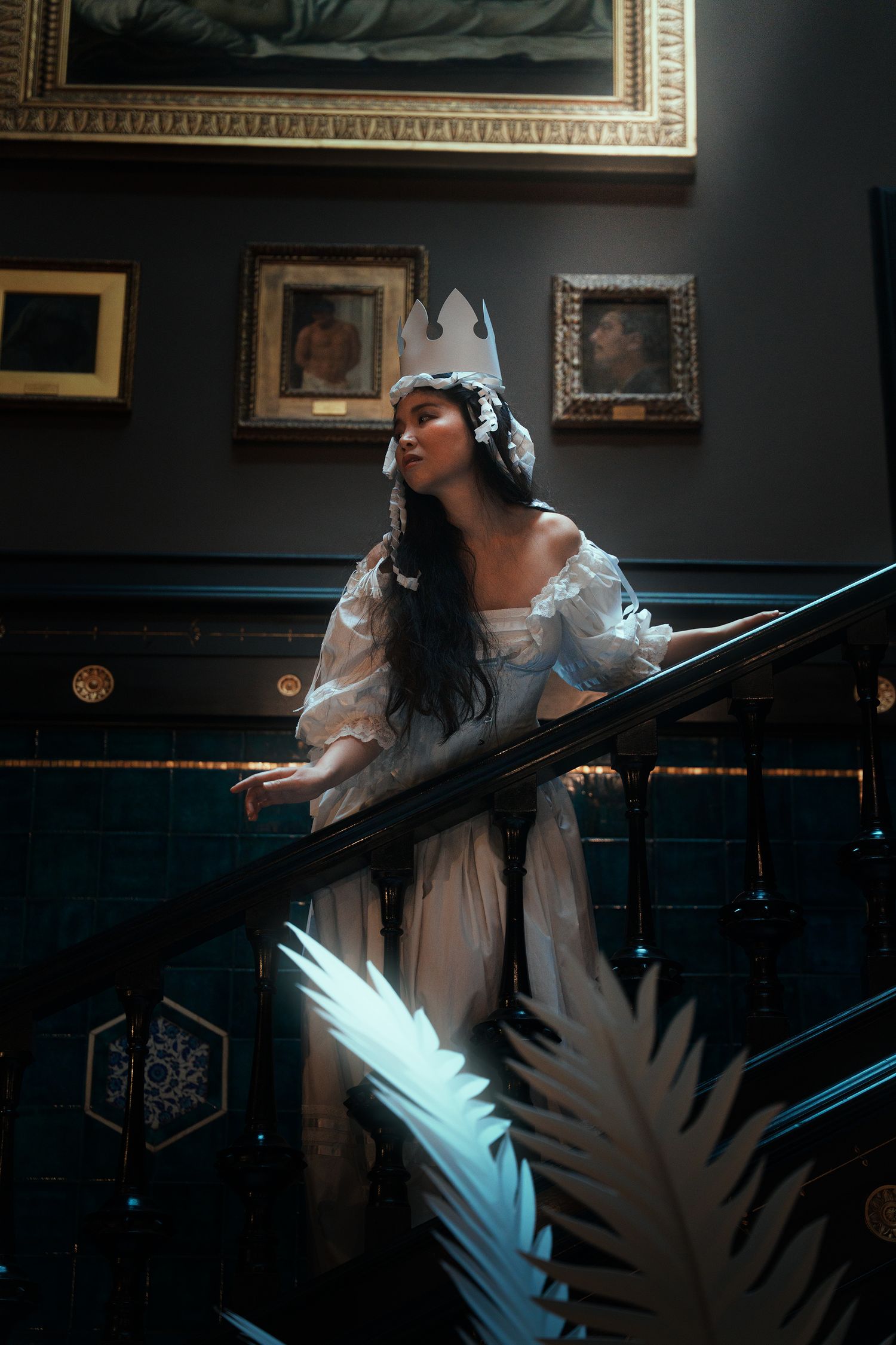 Elegant woman in white crown and gown on ornate dark staircase with framed paintings.