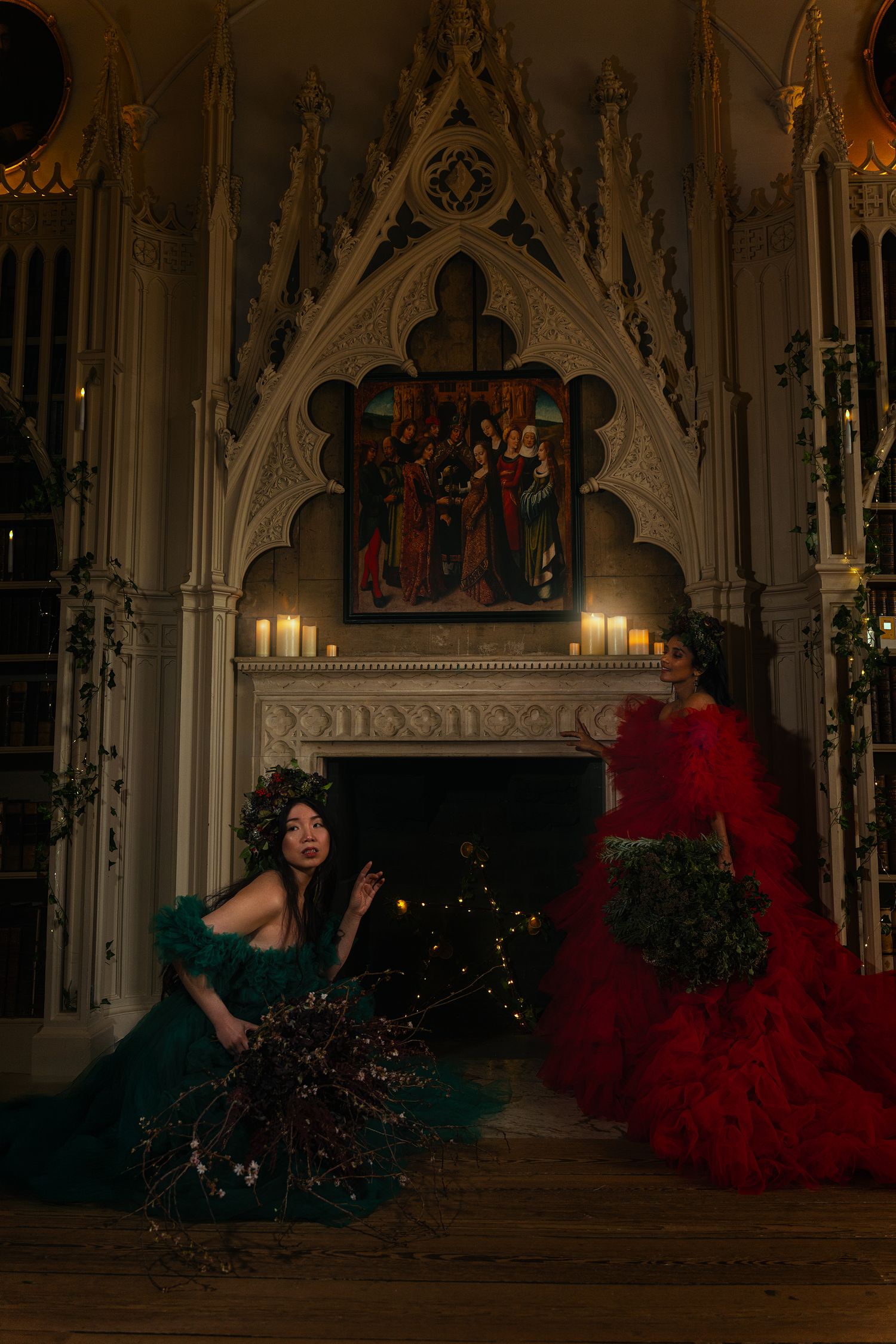 Two women in red and green gowns posed by Gothic fireplace with candles and Christmas decor.