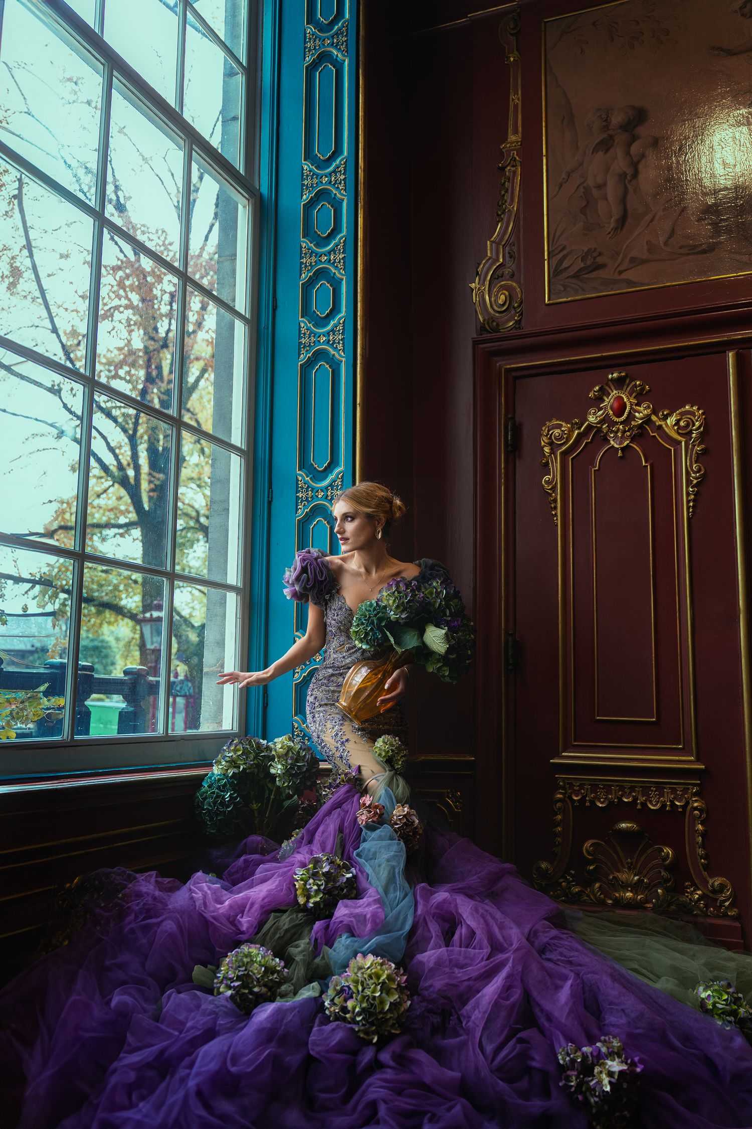A woman in a fantasy purple mermaid gown adorned with hydrangeas stands by a large window in an ornate room.