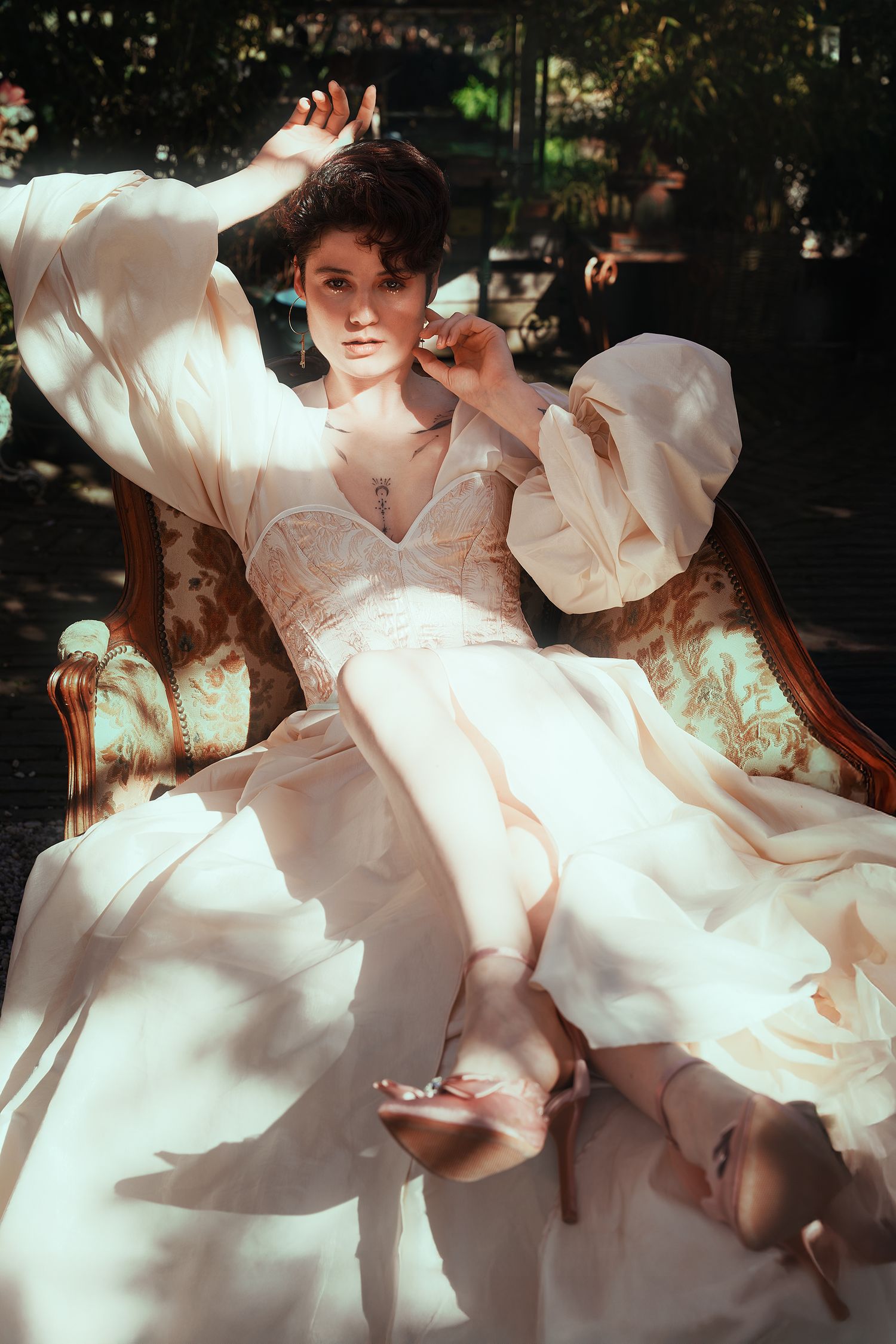 Woman in ivory gown with puff sleeves reclines in ornate chair bathed in dappled sunlight.