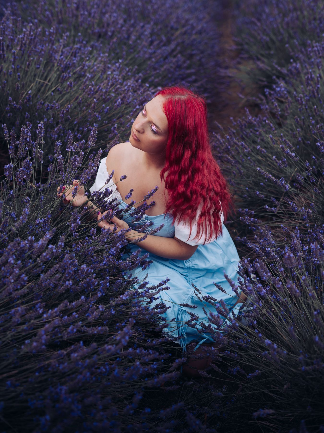 Woman with red hair in blue dress holding lavender bouquet in purple lavender field.