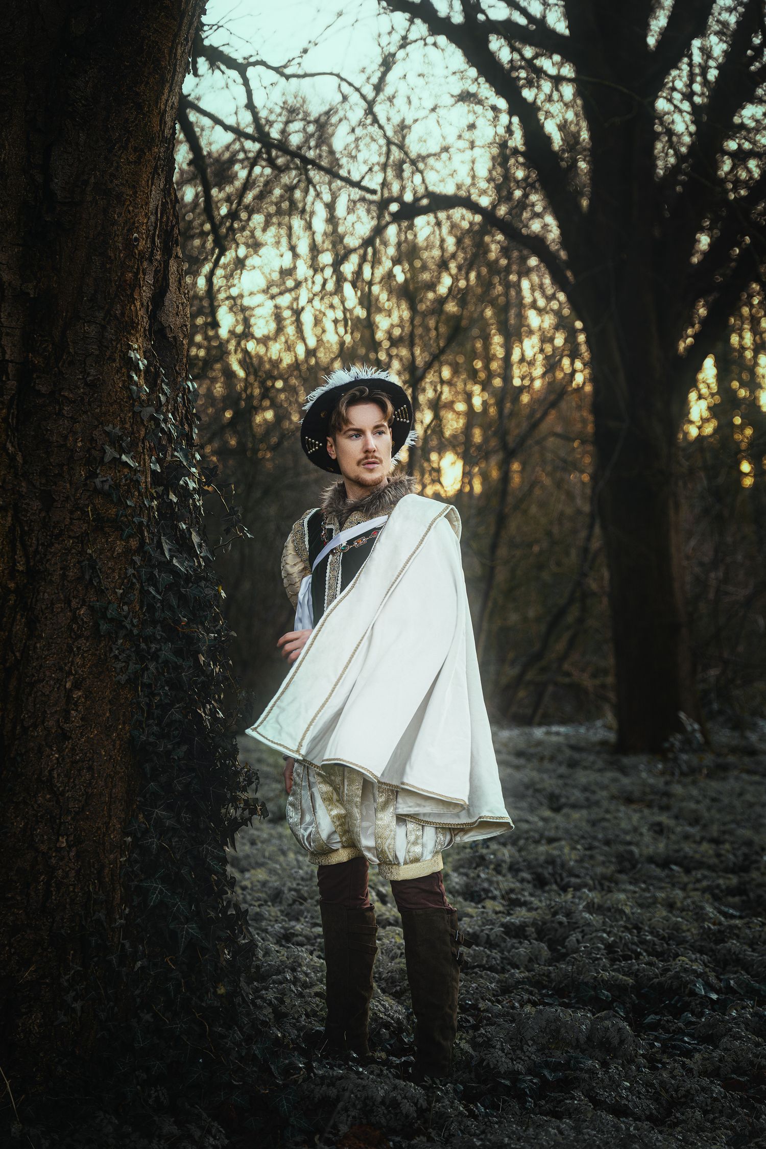 A man in Renaissance costume with white cape and hat stands in a dark autumn forest at sunset.