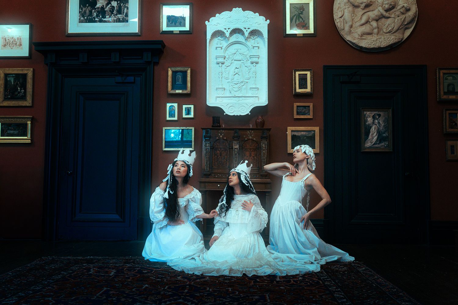Three women in white gowns and crowns posed in elegant red room with artwork.
