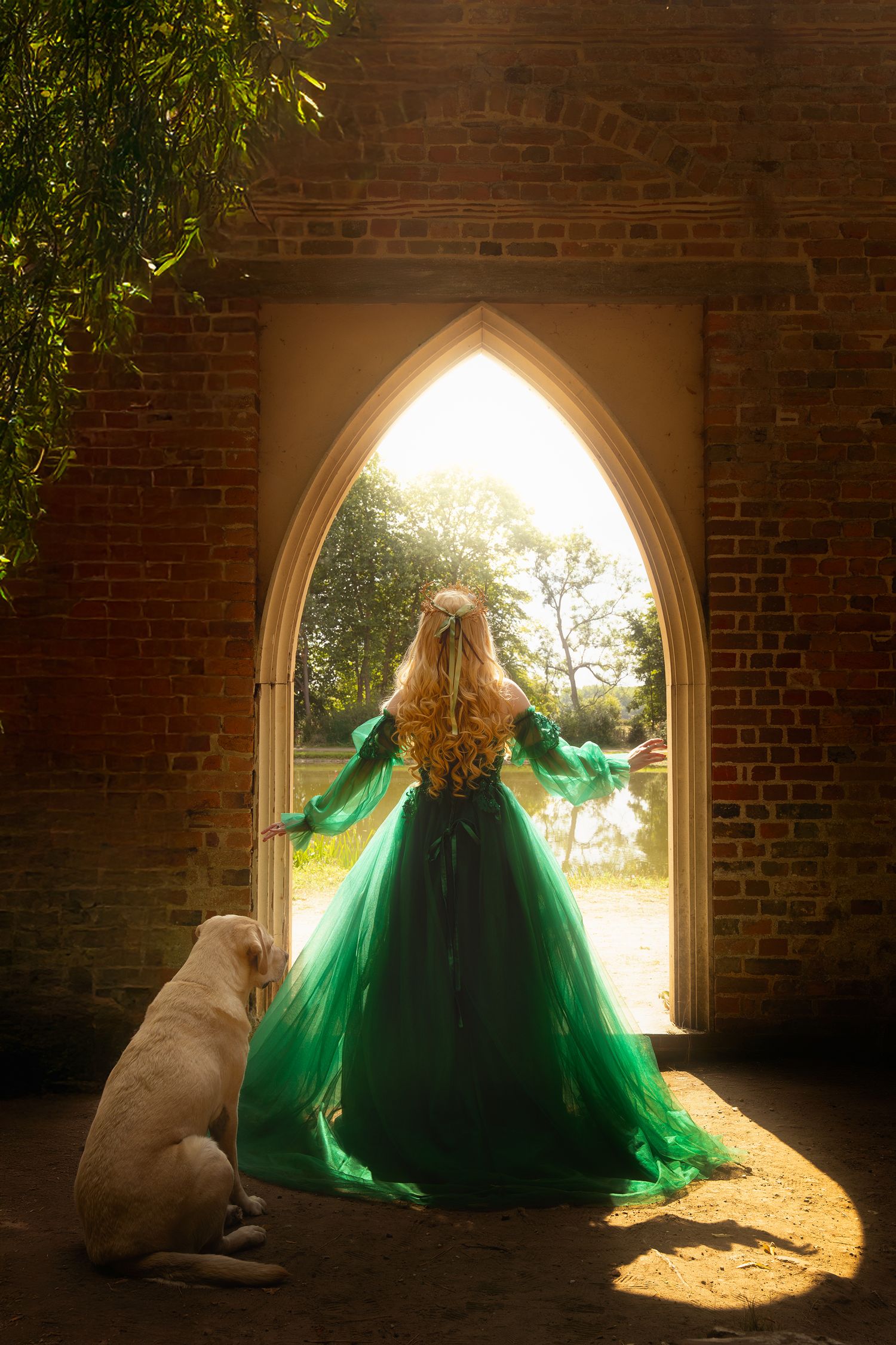 Woman in emerald green medieval gown standing in Gothic arch doorway with white dog.