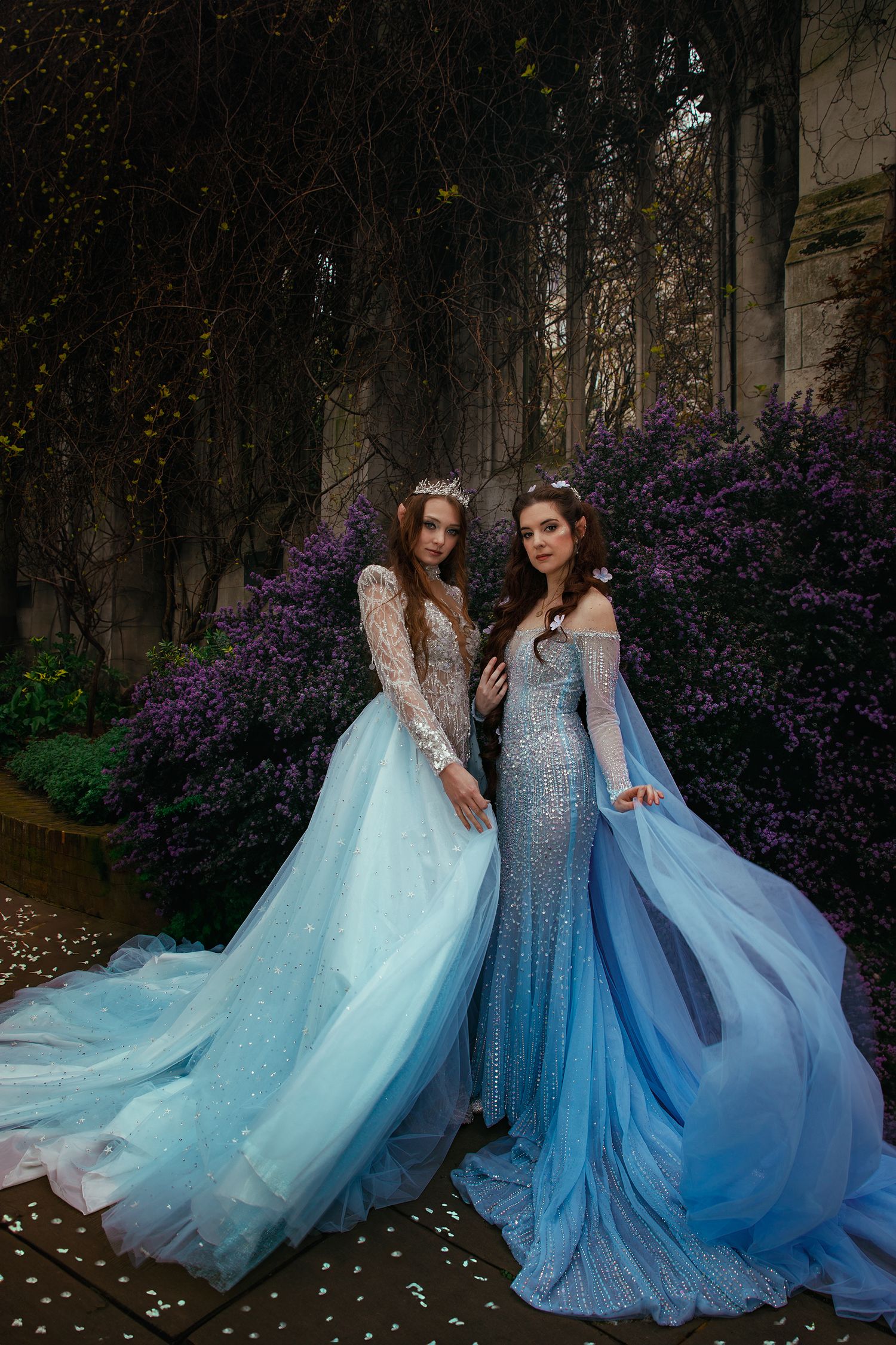 Two women in elegant blue fantasy gowns posing before flowering vines and stone wall.