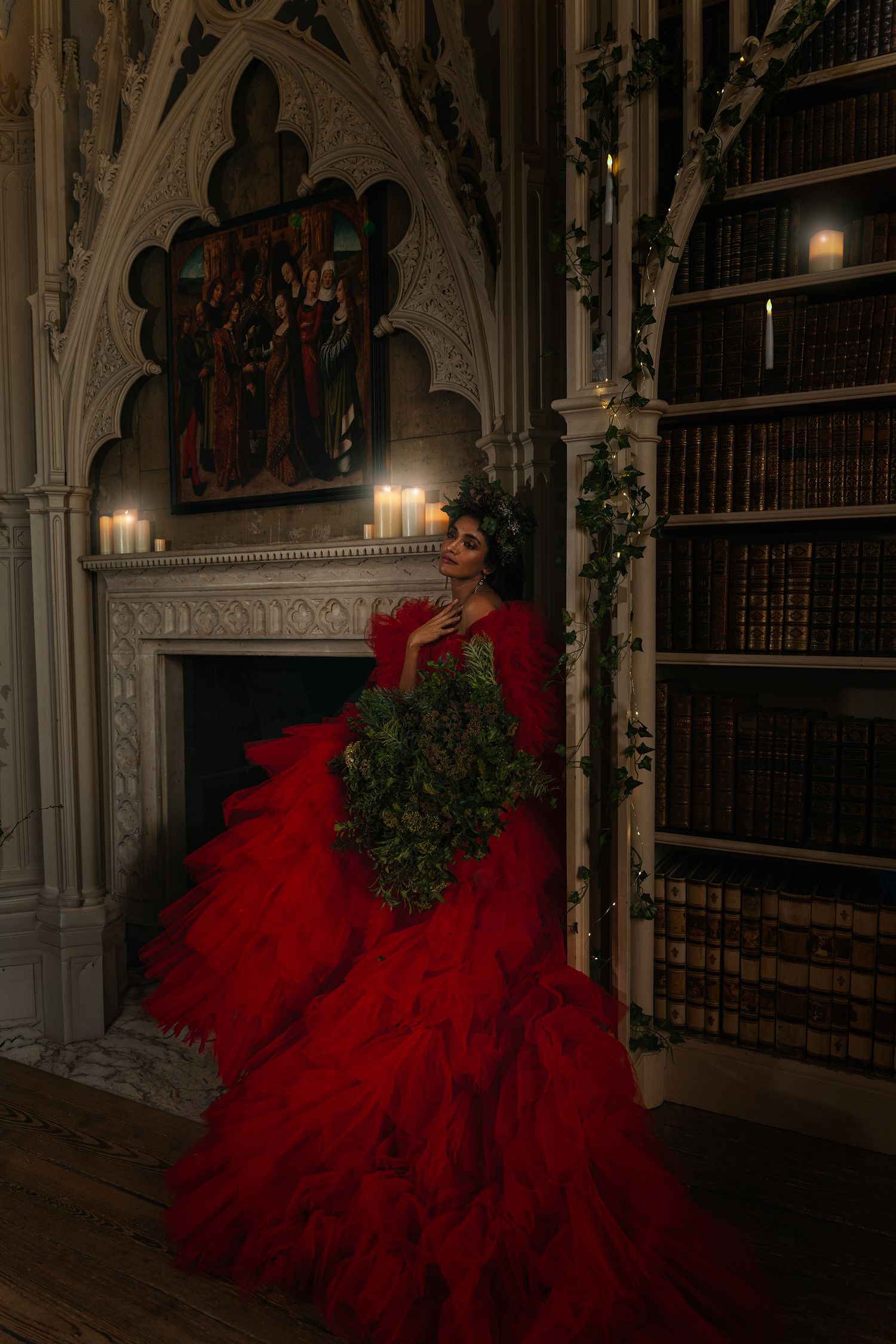 Woman in dramatic red ruffled ballgown holds greenery bouquet beside Gothic fireplace in candlelit library.