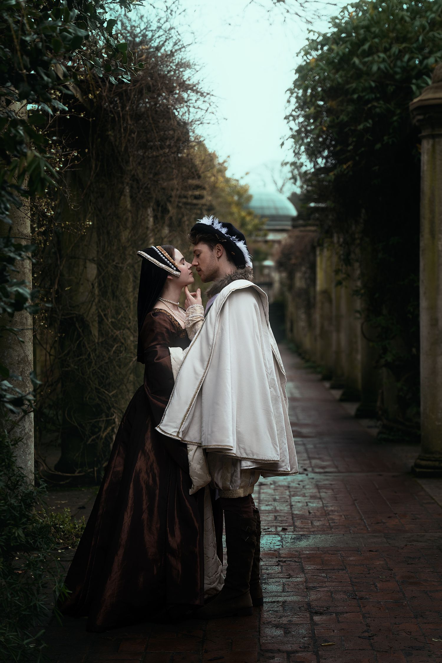 Medieval royal couple in historical costumes embracing in a misty stone garden corridor.