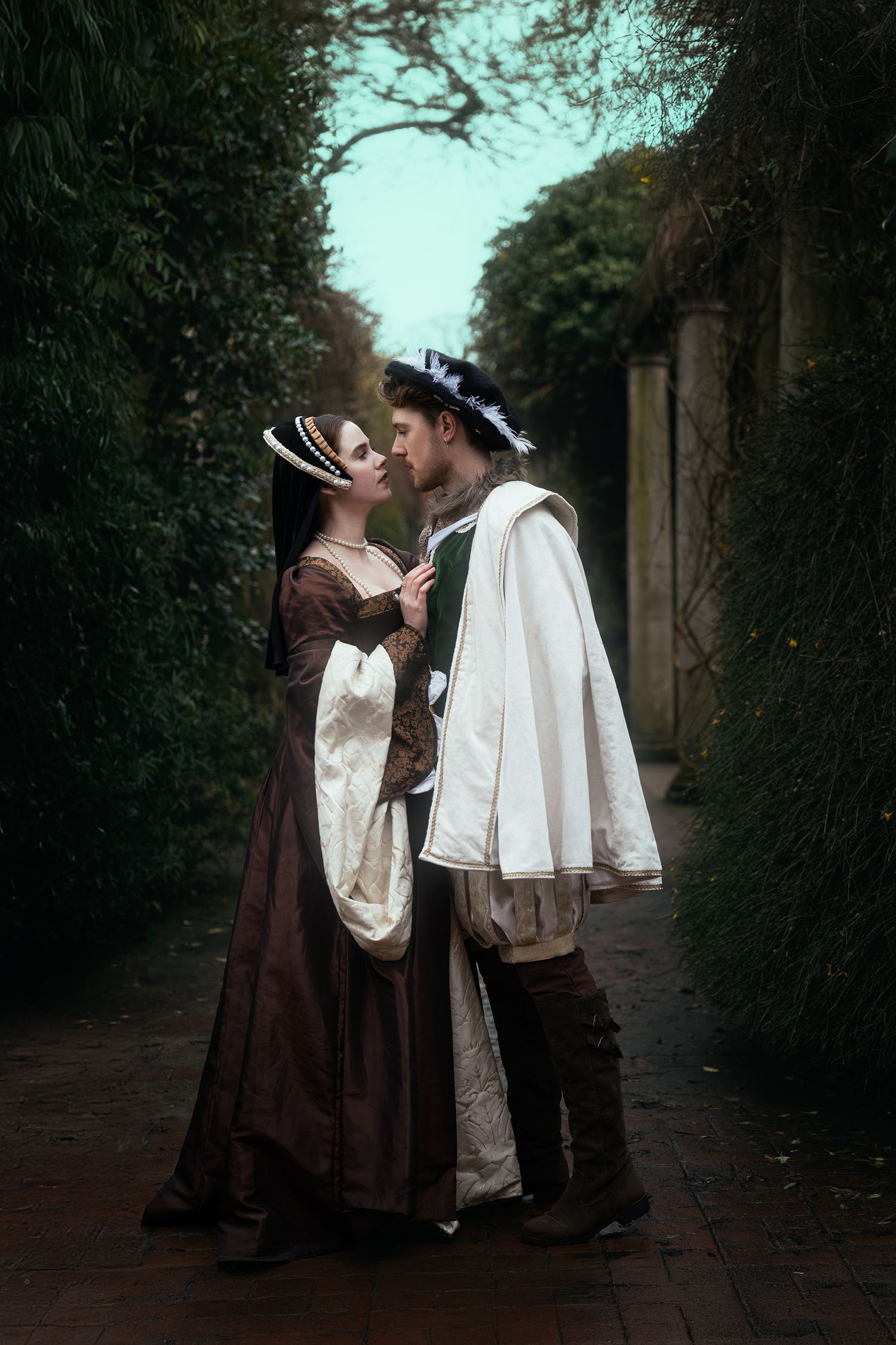 Renaissance couple in period costumes sharing a tender moment in a lush garden pathway.