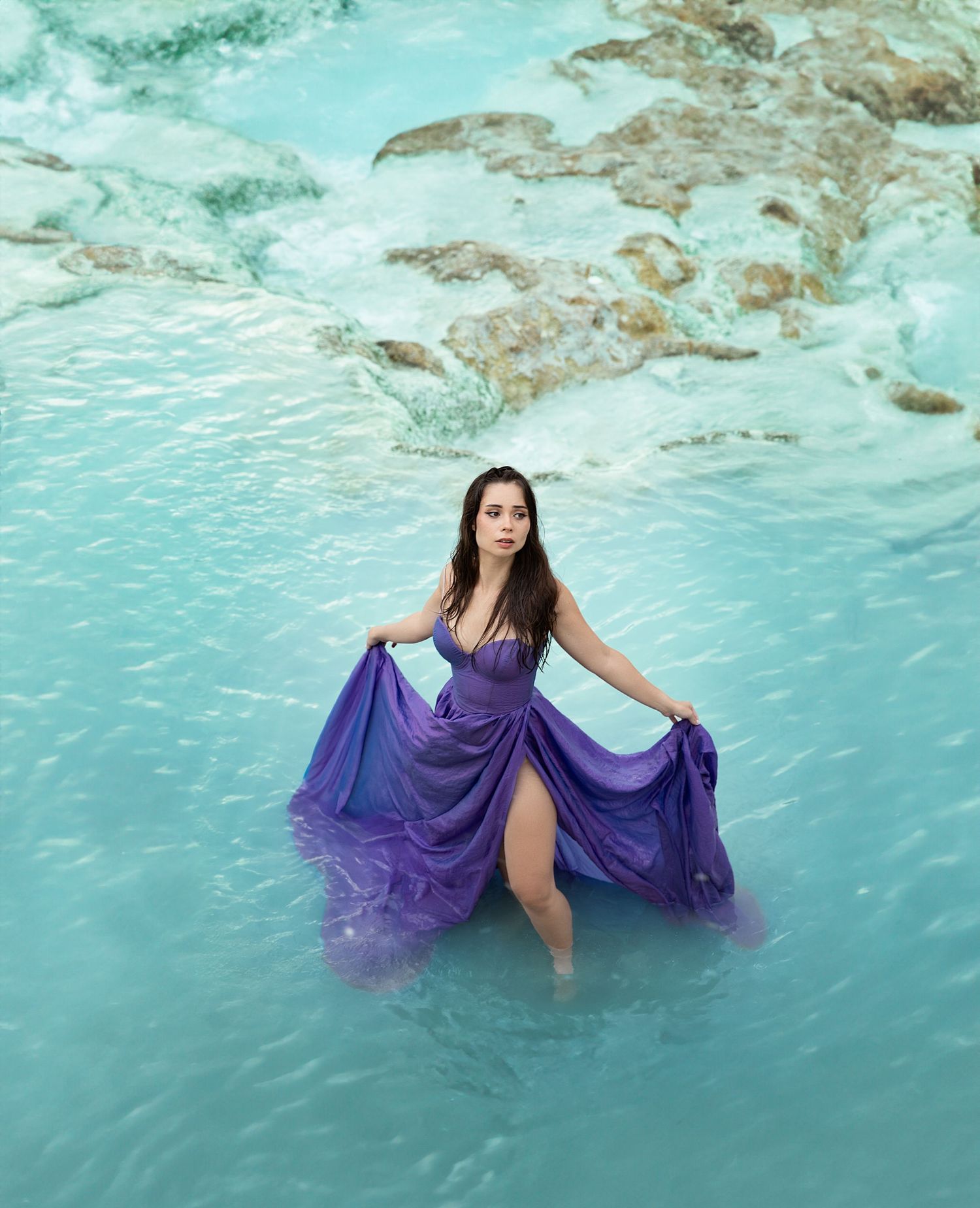 Woman in flowing purple gown standing in turquoise ocean waters with rocky shore backdrop.