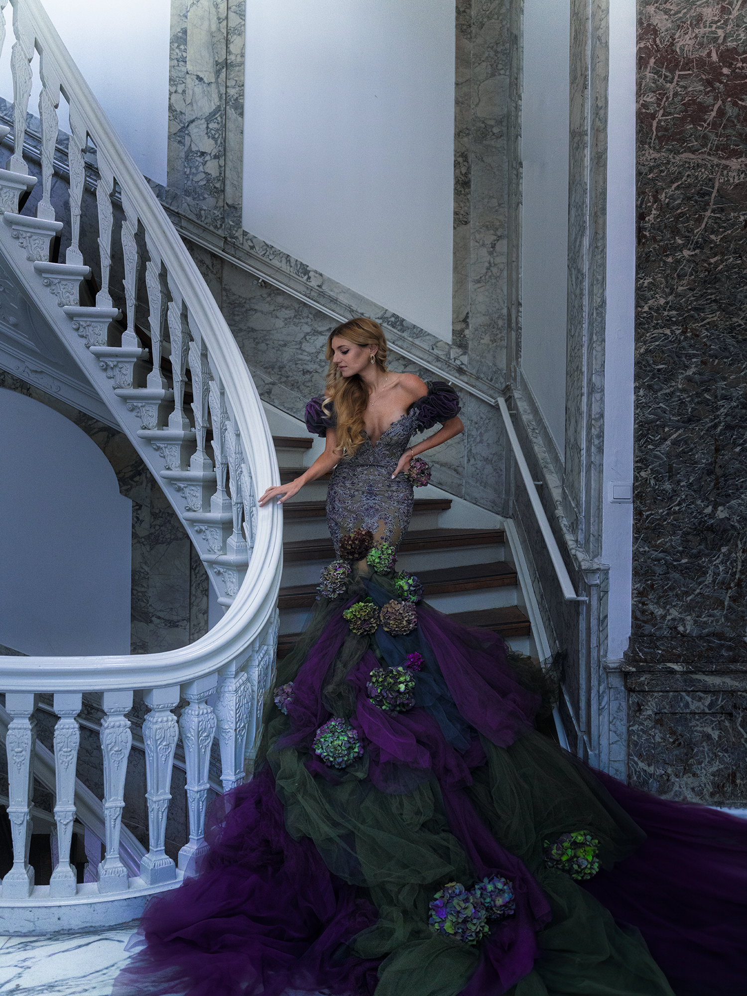 A woman in an elaborate purple and green gown with floral details poses gracefully on an ornate marble staircase.