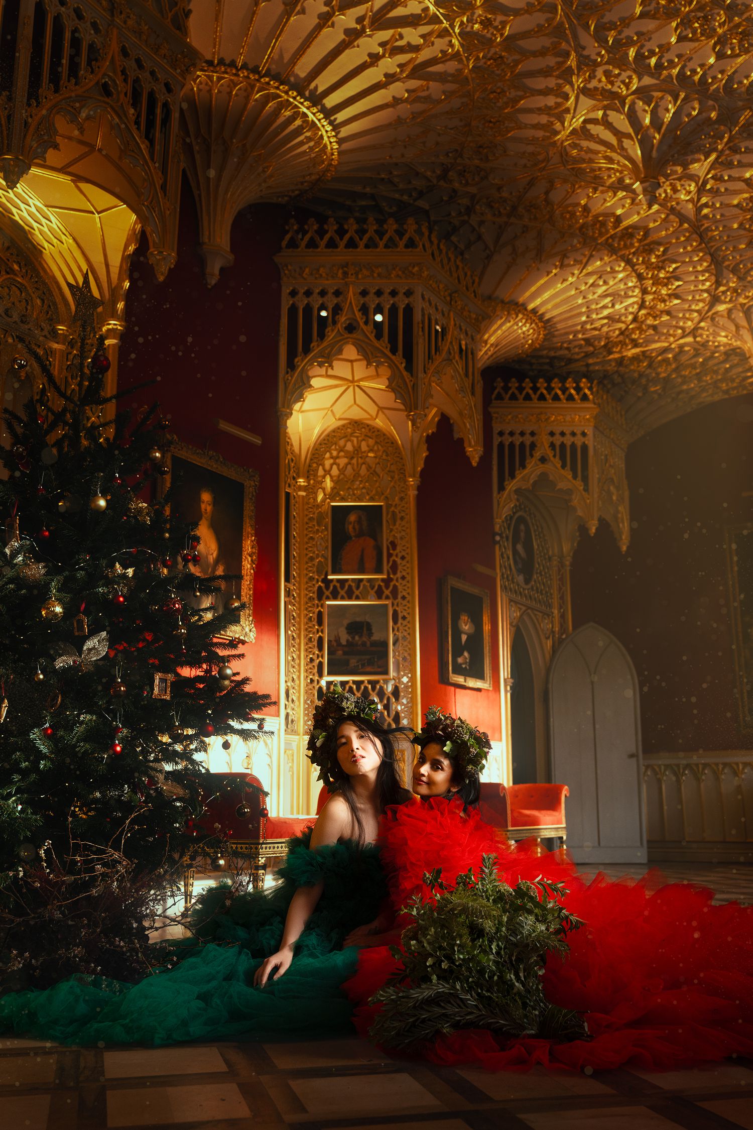 Two women in red and green gowns lying near Christmas tree in ornate golden Gothic interior.