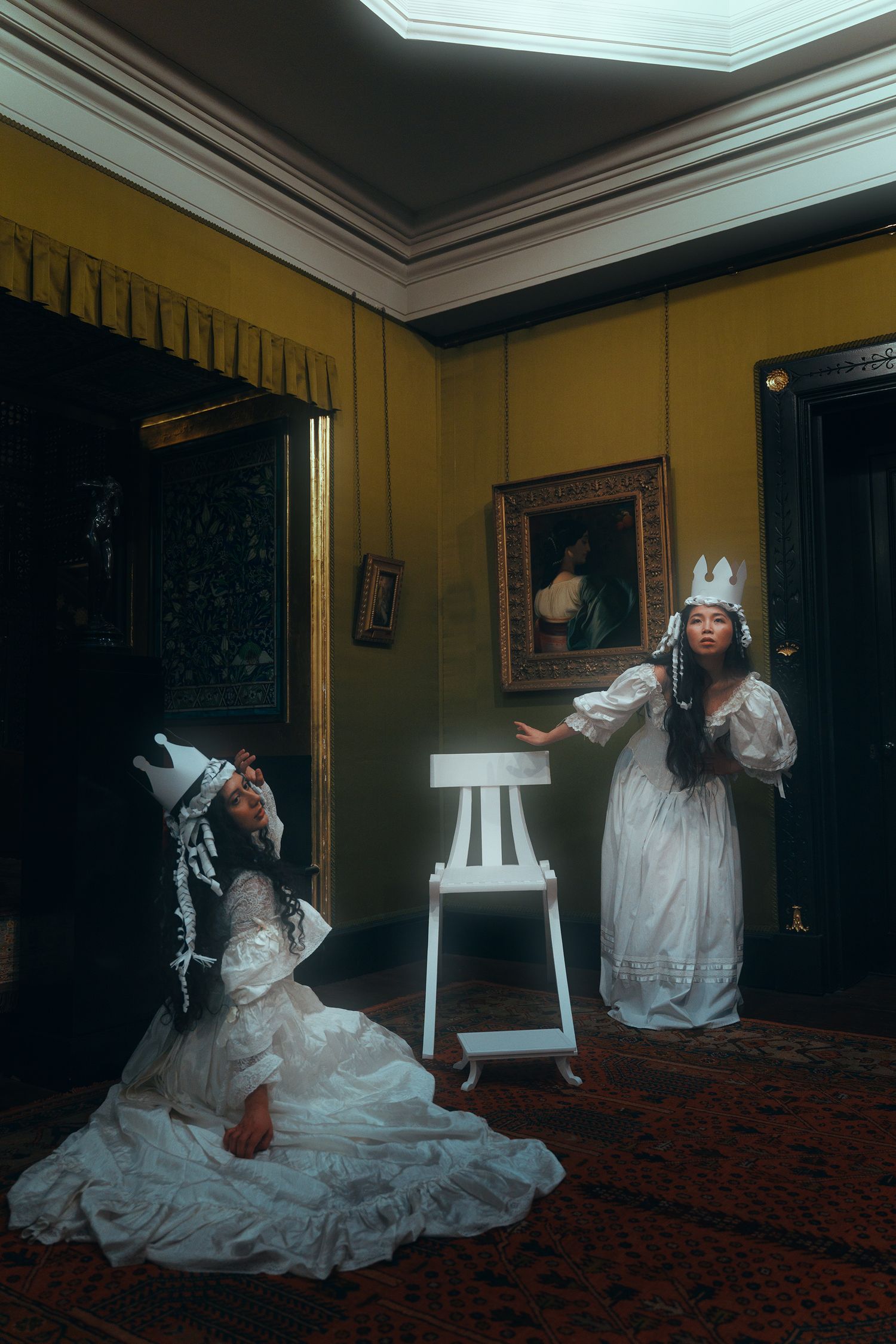 Two women in white Victorian gowns and paper crowns pose dramatically in an ornate yellow-walled room with classical paintings.