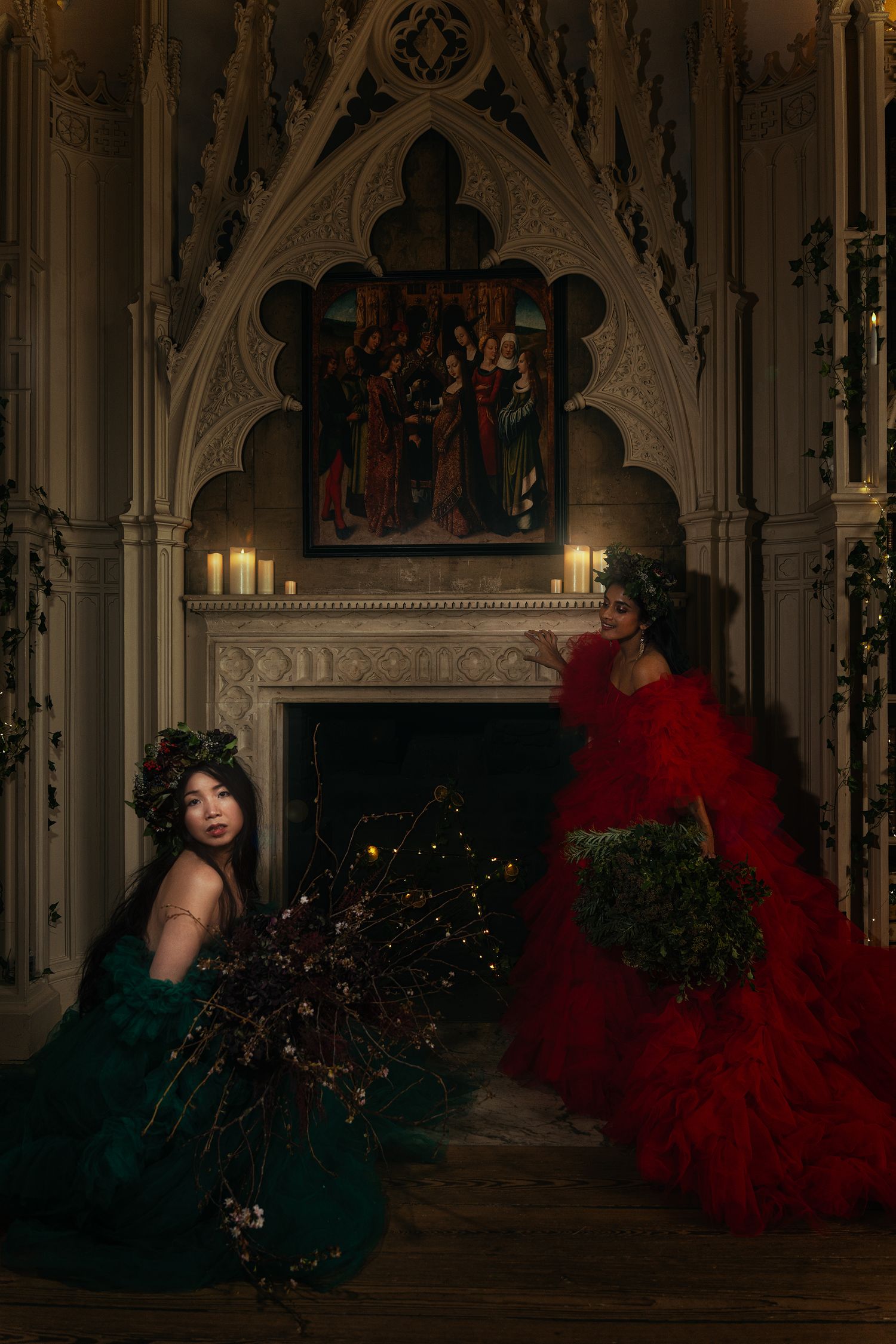 Two women in red and green ballgowns pose by an ornate Gothic fireplace with candles and a Renaissance painting.