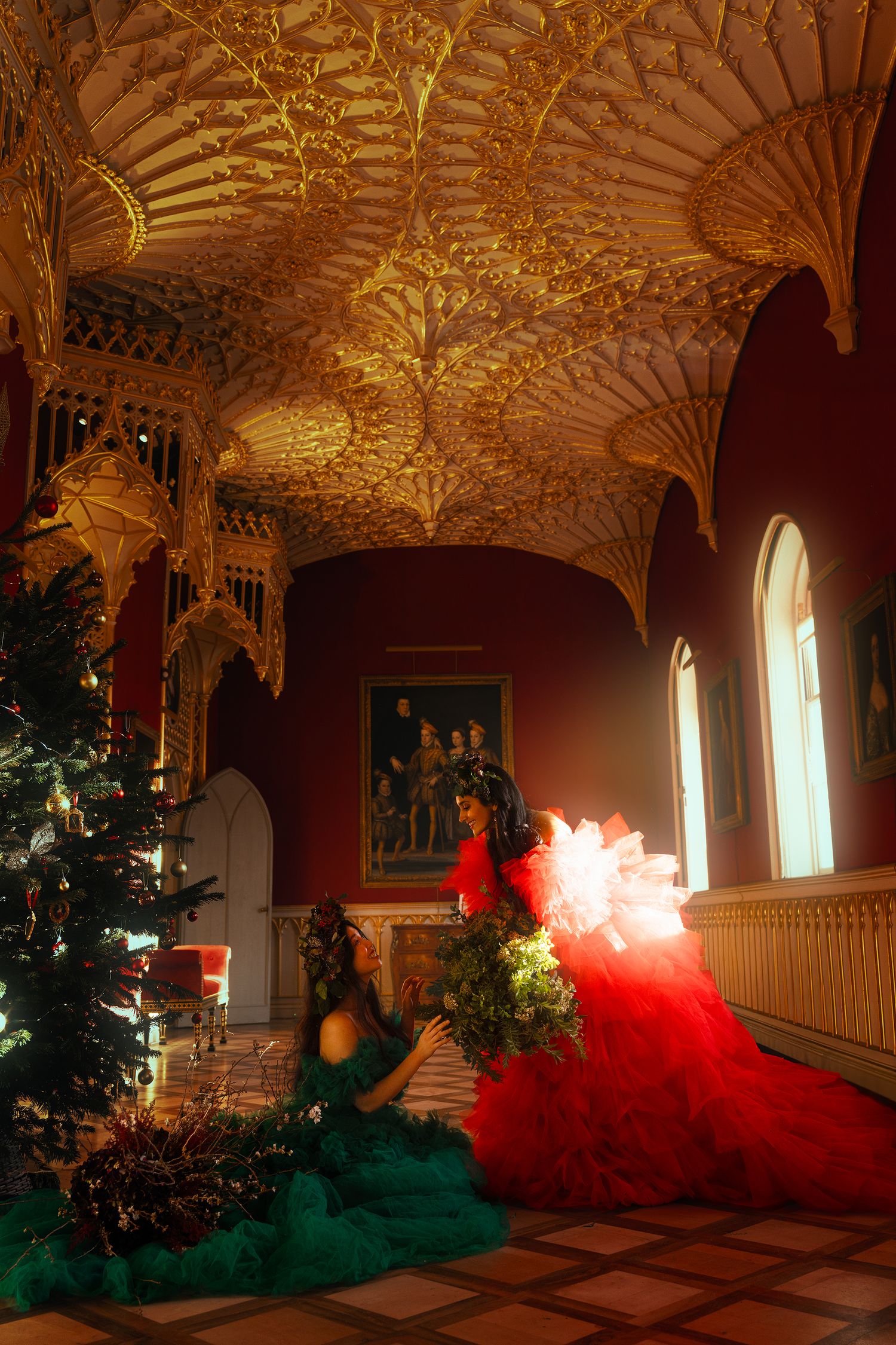 Two women in dramatic red and green gowns pose in an ornate golden Gothic palace room.